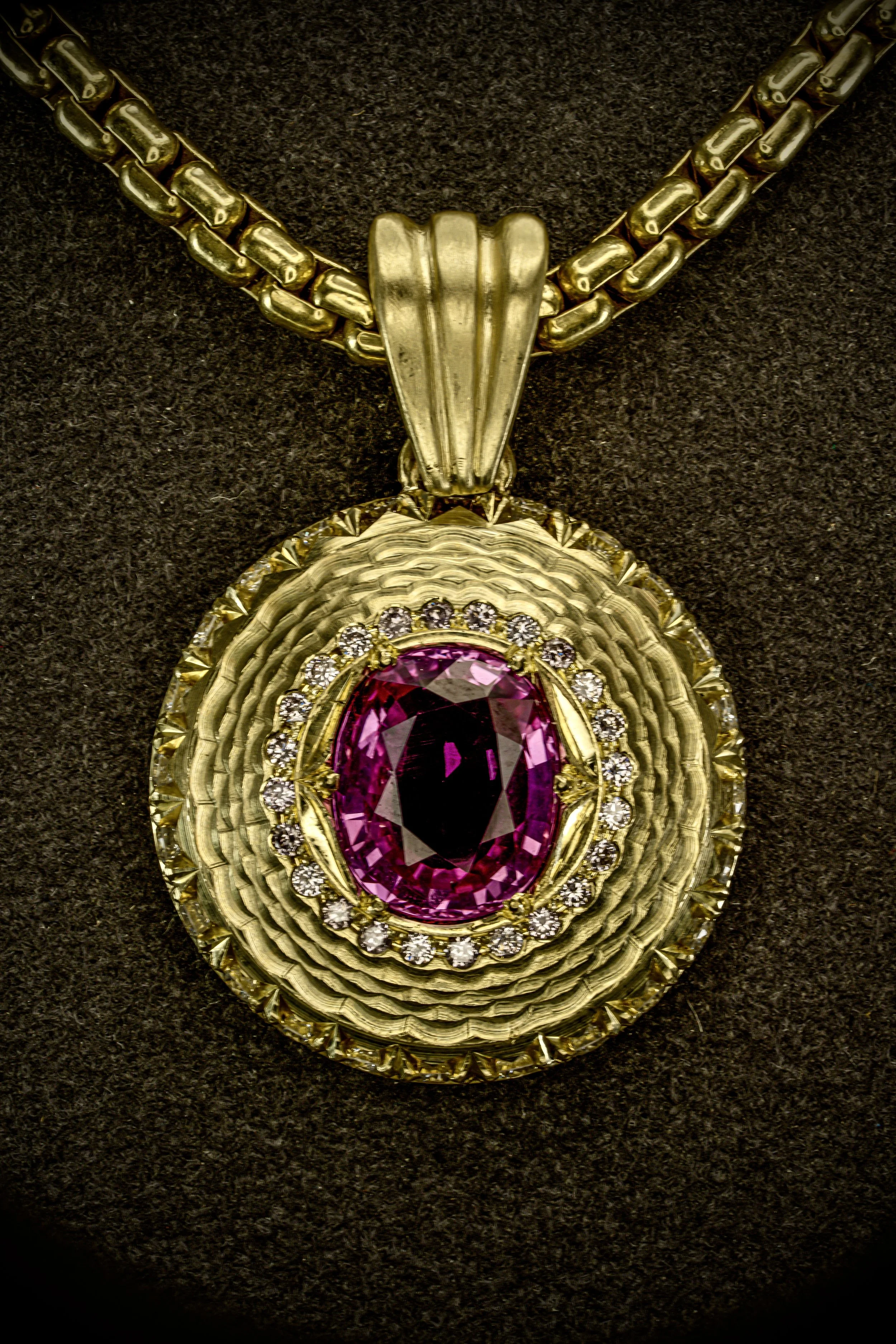 Gold medallion pendant with oval pink gemstone center, surrounded by a halo of small diamonds and layered textured gold detailing on a gold chain.