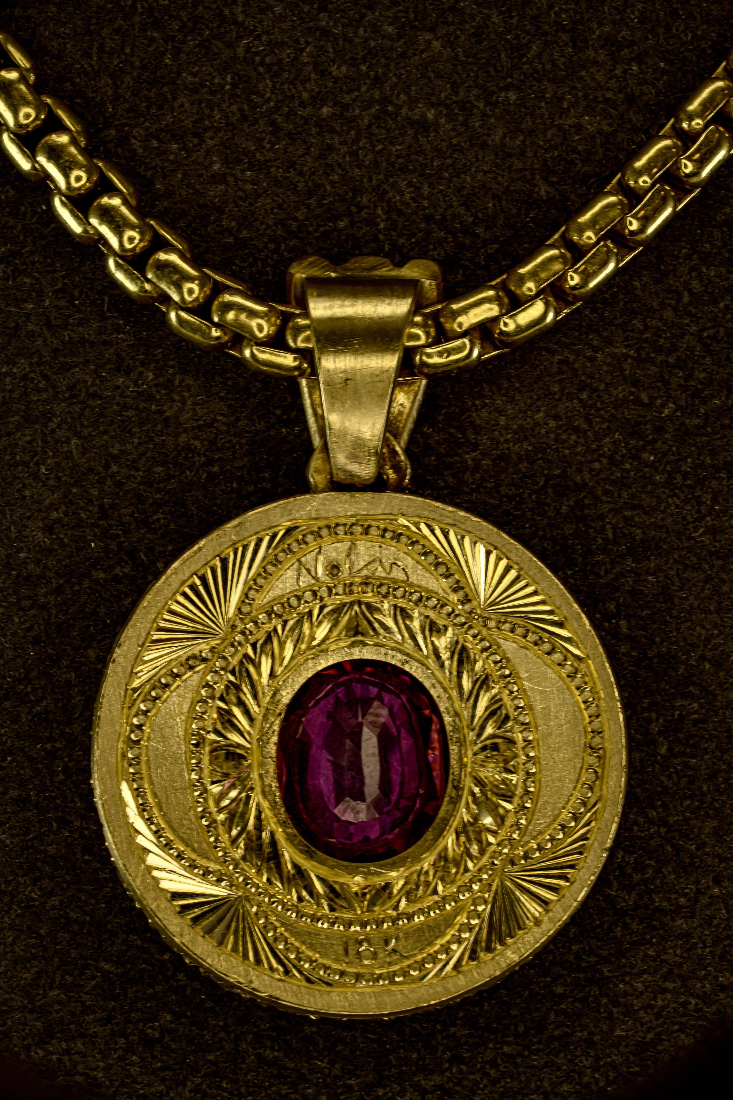 Reverse of gold medallion pendant showing engraved radial and geometric patterns with an oval pink gemstone visible in the center, suspended from a gold chain.