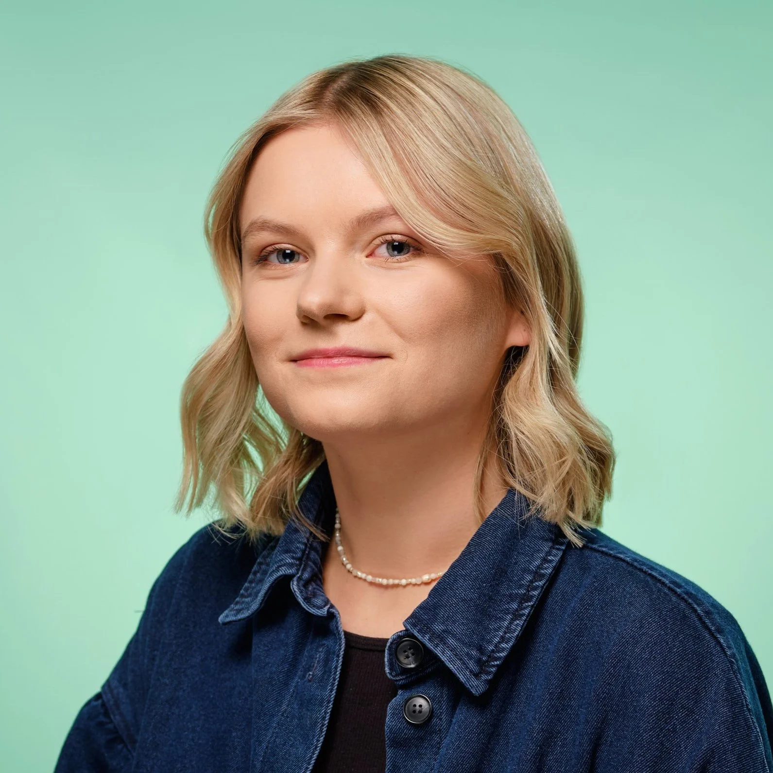 Portret kobiety o blond włosach i niebieskich oczach, ubranej w dżinsową kurtkę nałożoną na czarny top i naszyjnik z pereł, na zielonym tle.