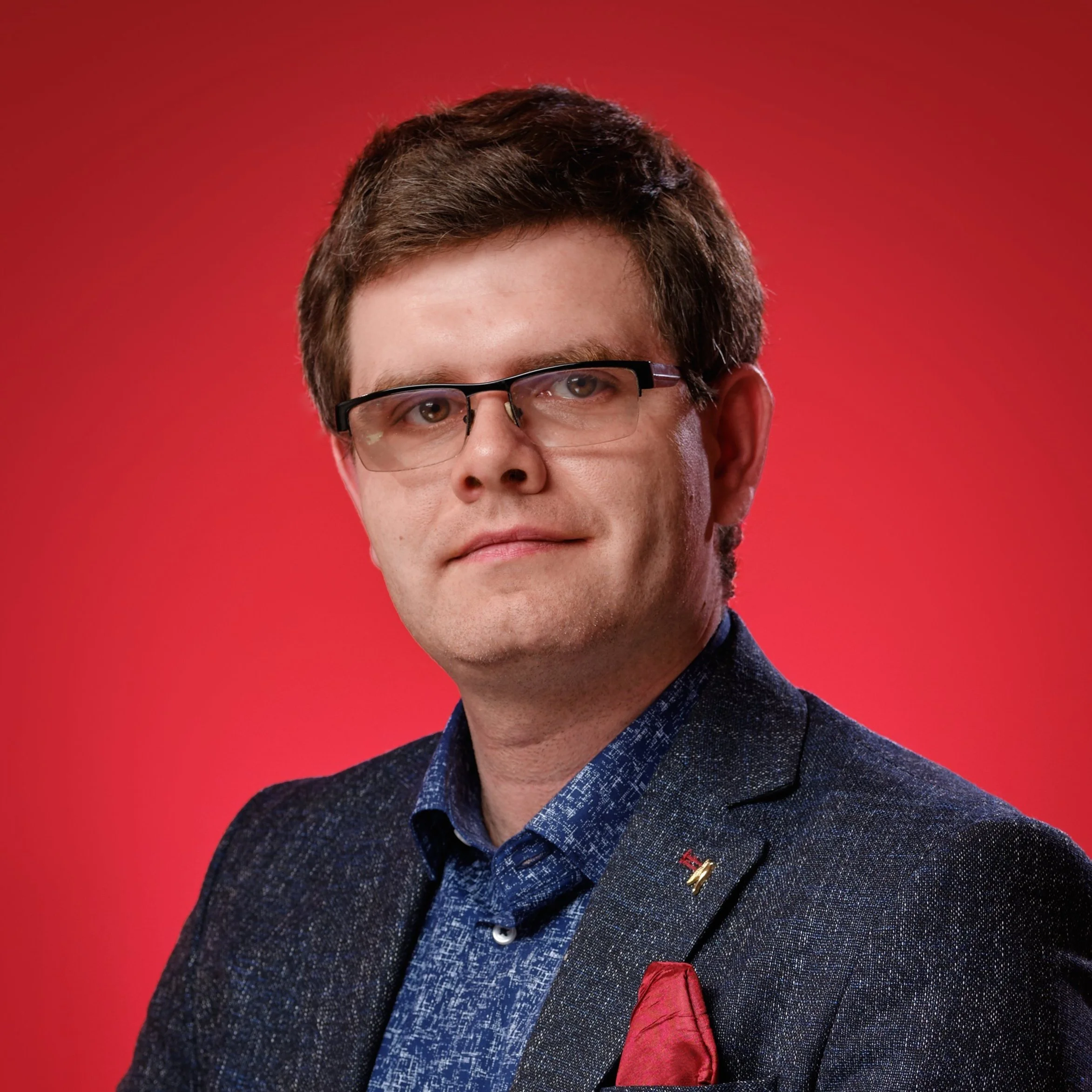 Zdjęcie z bliska przedstawiające mężczyznę z krótkimi brązowymi włosami i okularami, ubranego w granatową marynarkę i niebieską koszulę na jednolitym czerwonym tle.