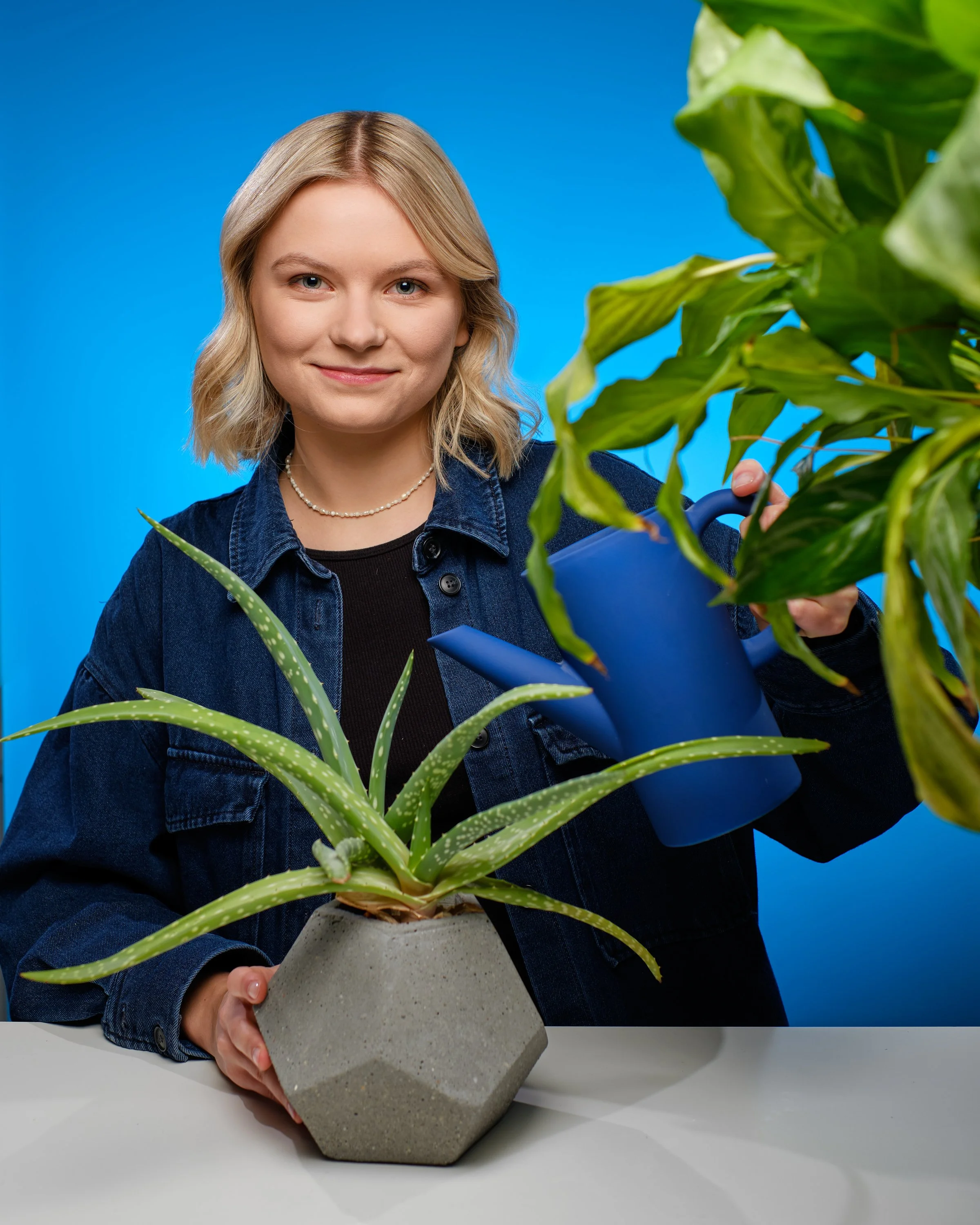 Młoda kobieta o blond włosach i niebieskich oczach trzyma doniczkową roślinę aloesu w szarej, geometrycznej doniczce na niebieskim tle.
