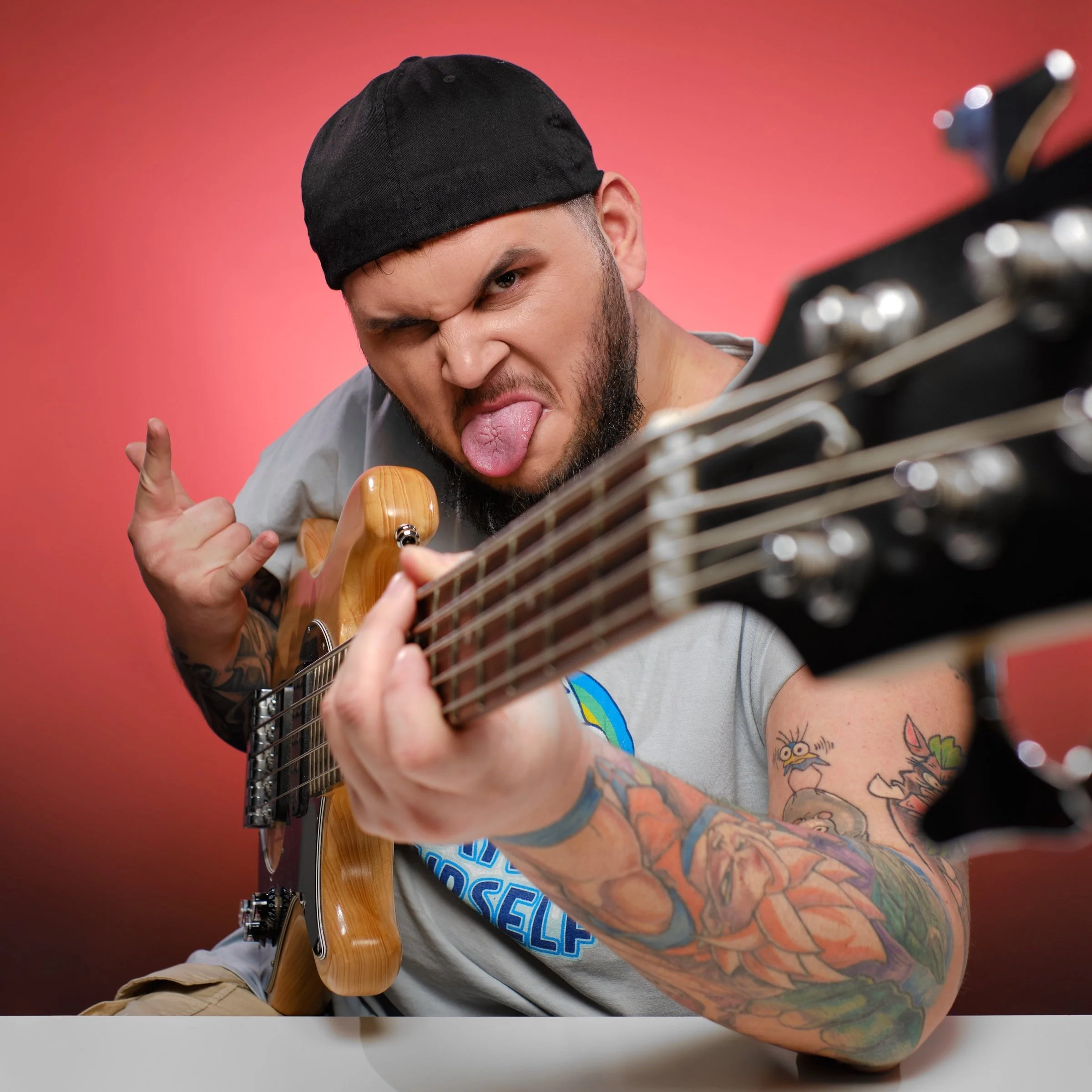 Wytatuowany mężczyzna z brodą i tatuażami na ramieniu gra na gitarze elektrycznej. Ma na sobie szary T-shirt, czarną czapkę z daszkiem skierowaną do tyłu i robi figlarną minę, wysuwając język na różowym tle.