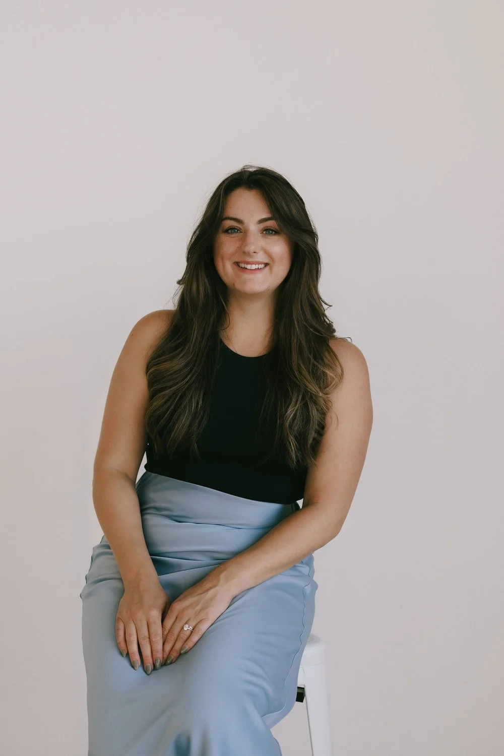 A woman with long dark hair, wearing a black sleeveless top and light blue satin skirt, smiling while sitting on a white stool against a plain white background.
