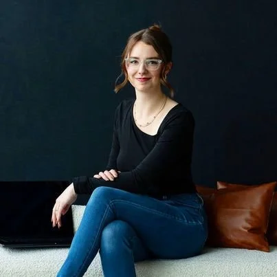 A woman with glasses sitting on a couch with dark background, smiling at the camera.