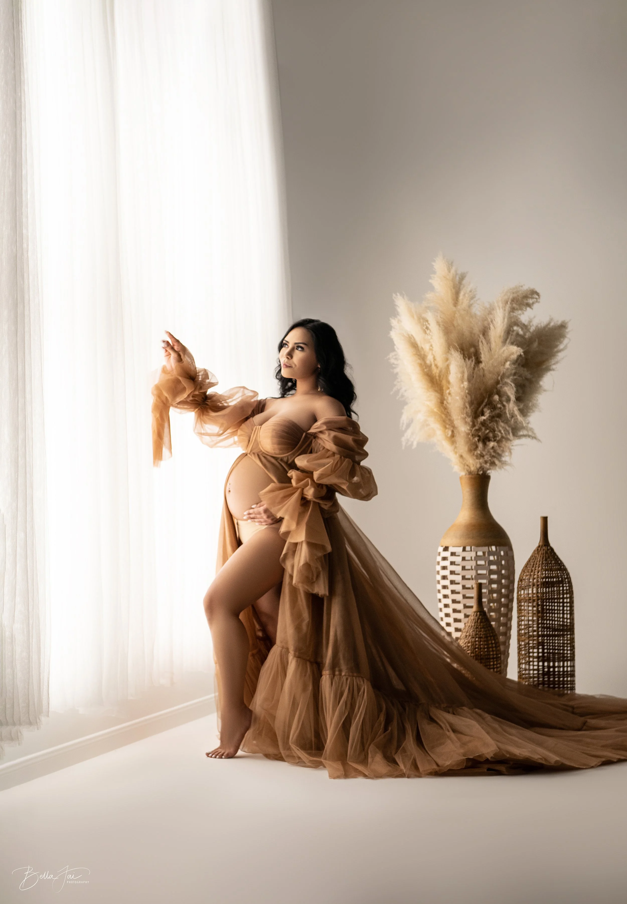 Pregnant woman in beige sheer gown standing by a window with white curtains, with large pampas grass and woven vases in the background.