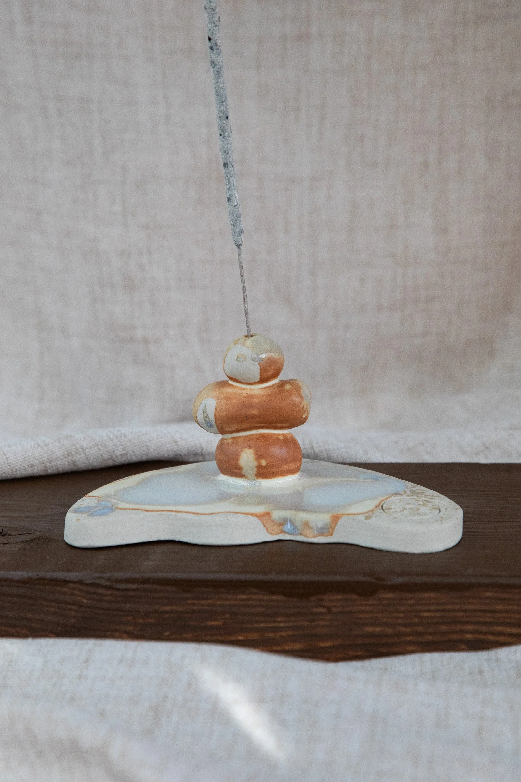 A ceramic sculpture with stacked rounded shapes in beige, orange, and cream, on a wooden surface with a textured beige background.
