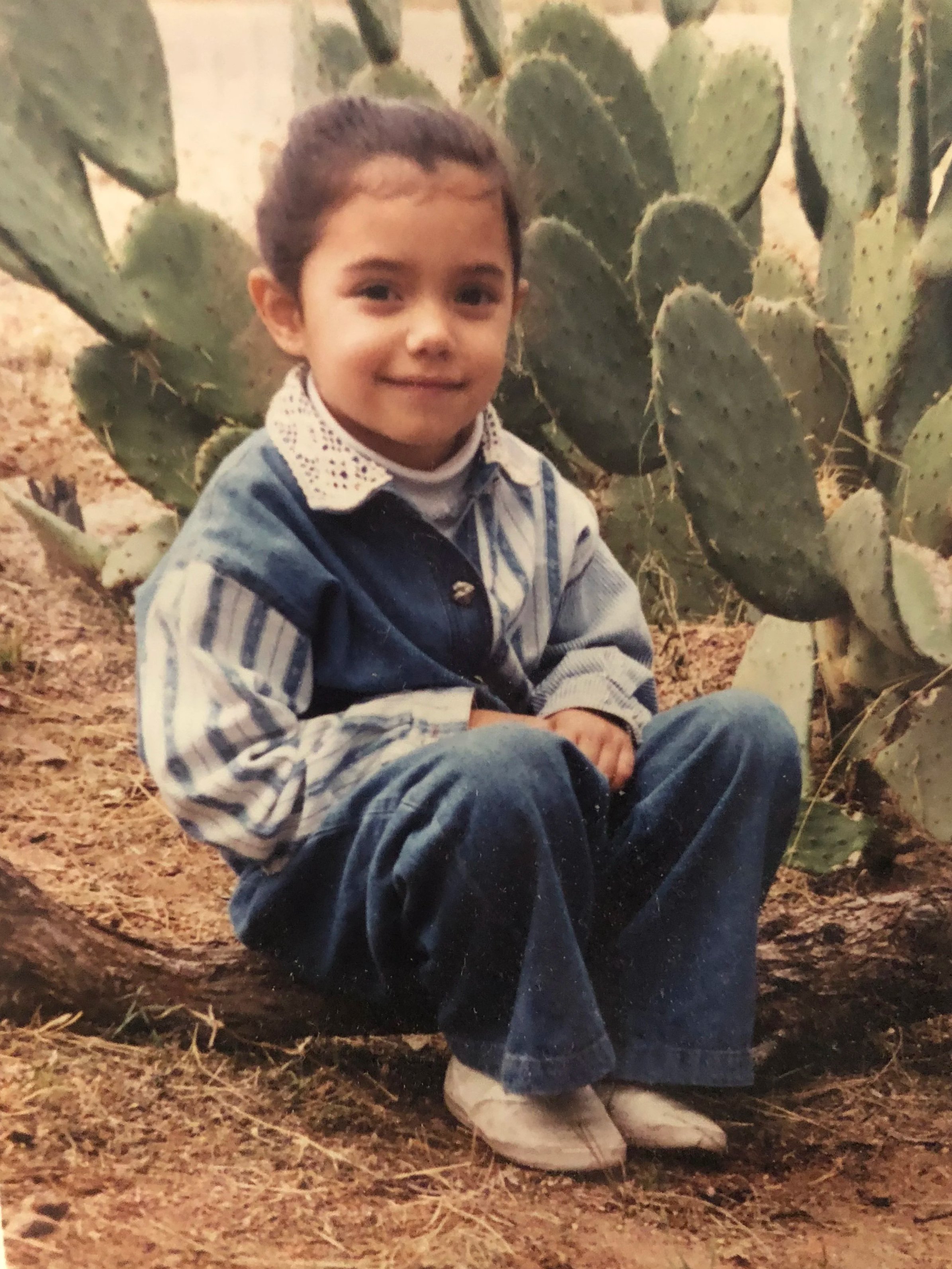 A young girl sitting on the ground in front of a large prickly pear cactus. She is wearing a blue and white striped shirt with a white collar, a denim vest, and light-colored shoes. The girl has her hair tied back and is smiling at the camera.