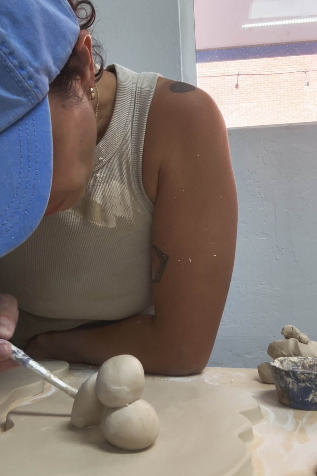 A person sculpting clay in a workshop, wearing a blue cap and a beige tank top, with dust and small clay pieces on their arm and workspace.