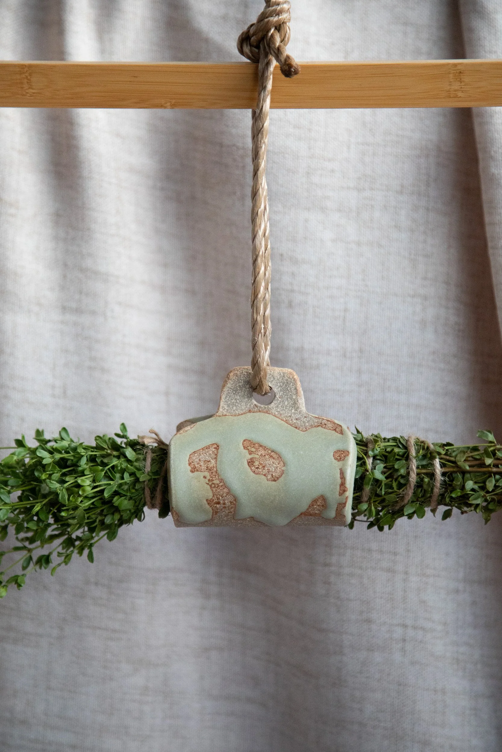 A hanging decorative piece with a ceramic soap dispenser mounted on a lush green faux garden, suspended by a thick, natural fiber rope tied to a wooden dowel, set against a neutral fabric backdrop.