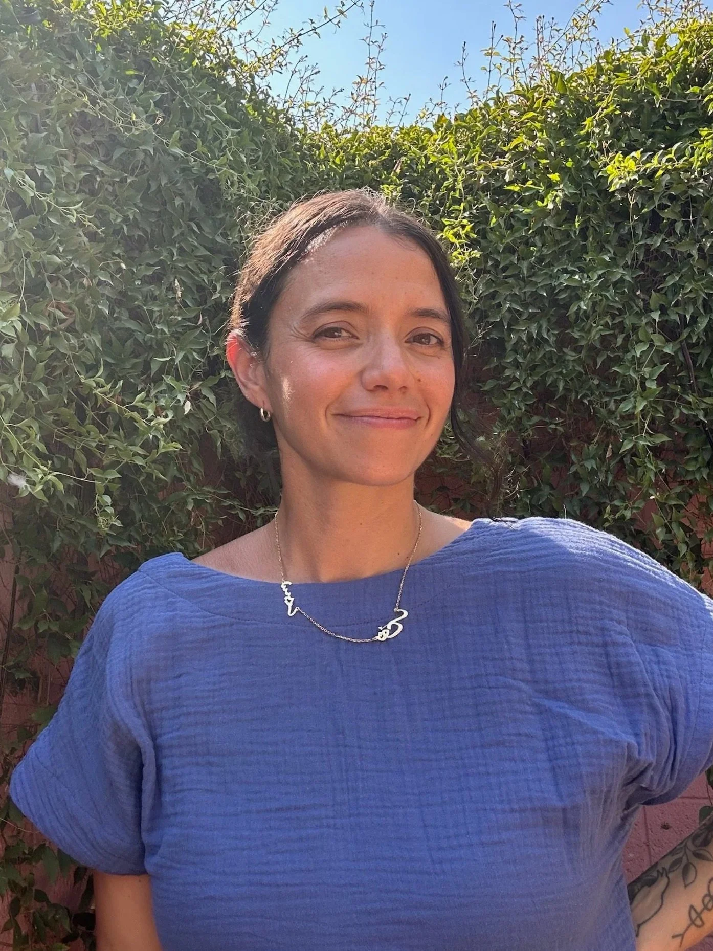 A woman in a blue top with a necklace, standing outdoors in front of green foliage under a clear blue sky, smiling at the camera.