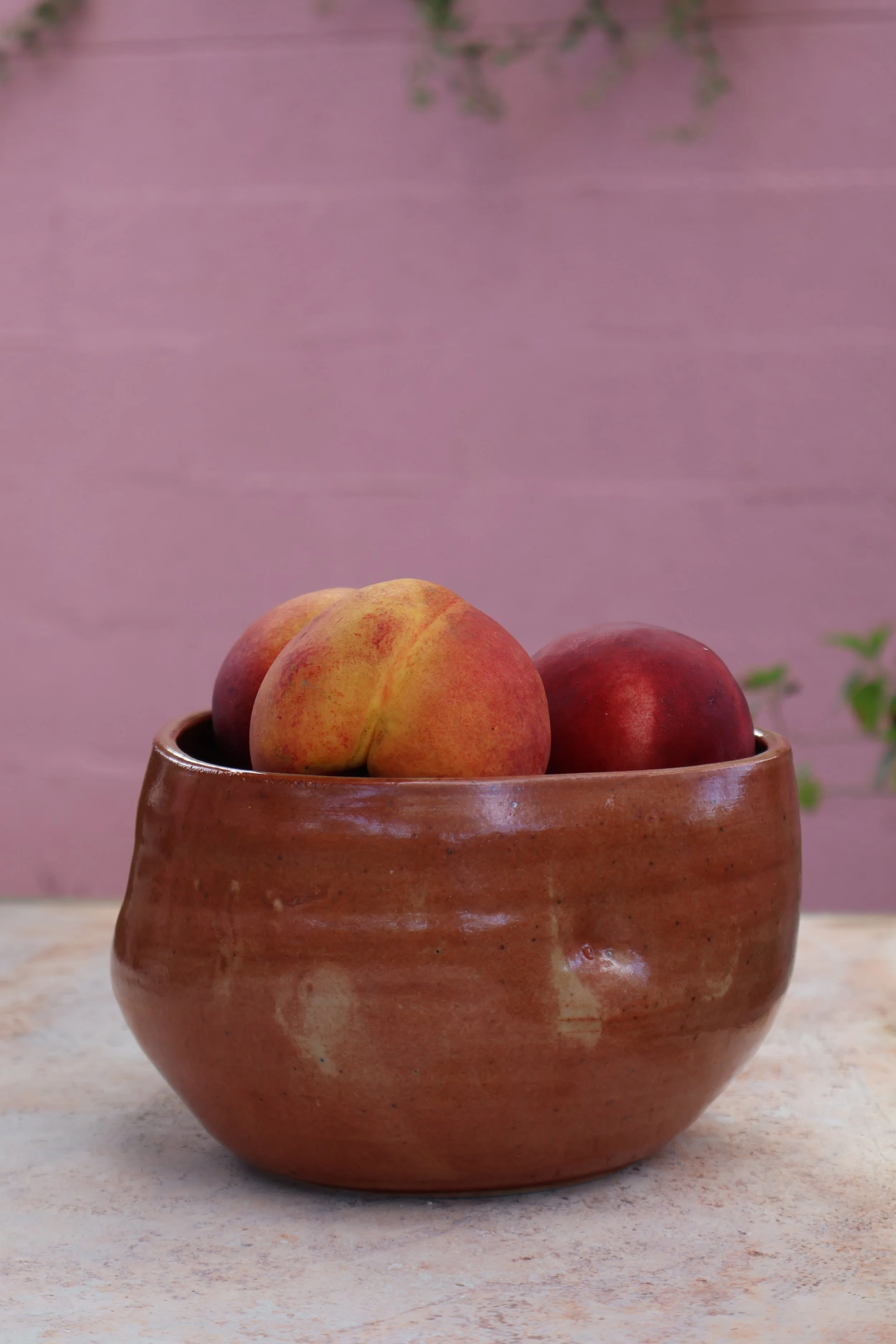 A ceramic bowl with three peaches placed on it, set on a beige surface against a pink wall background.
