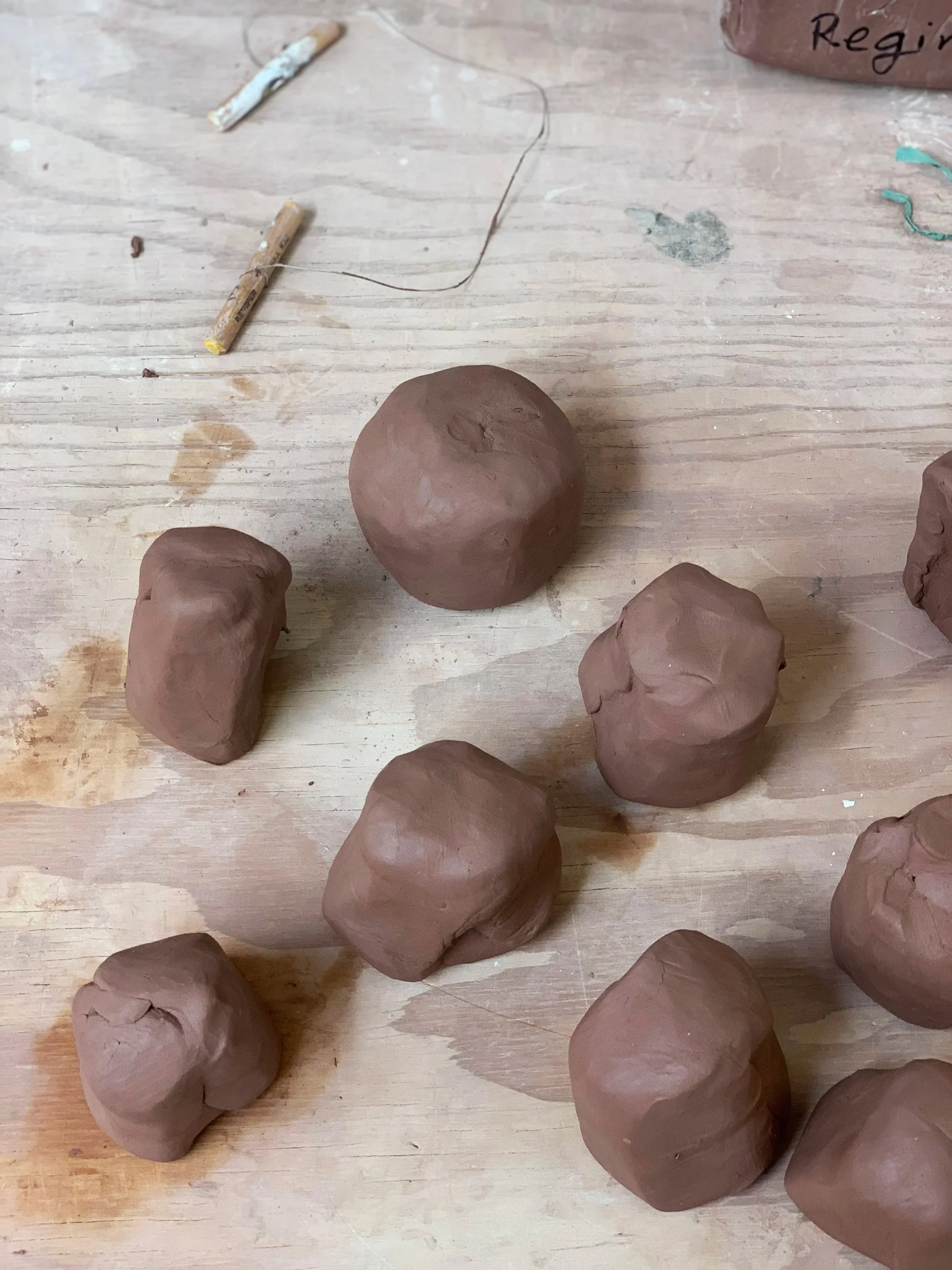 Several unfinished clay sculptures and clay tools on a wooden table, with some writing on the clay in the background.