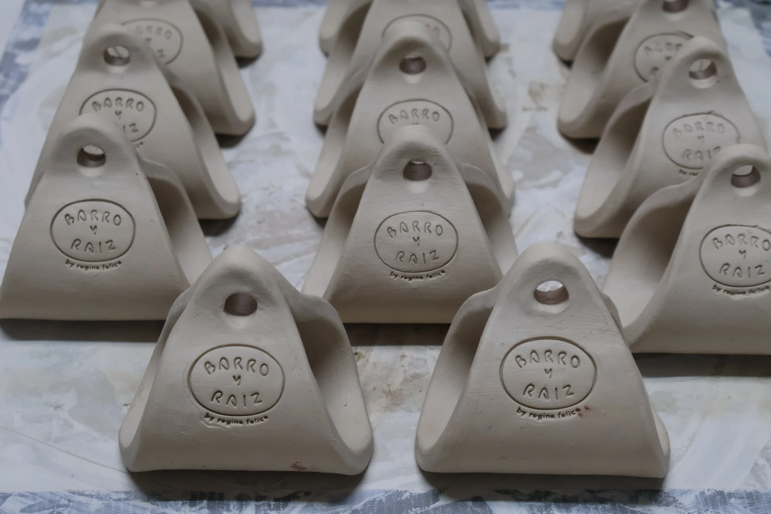 Multiple white ceramic satchel forms arranged in rows on a surface, each stamped with the words 'BARRO y RAIZ' and 'by regina felice'.