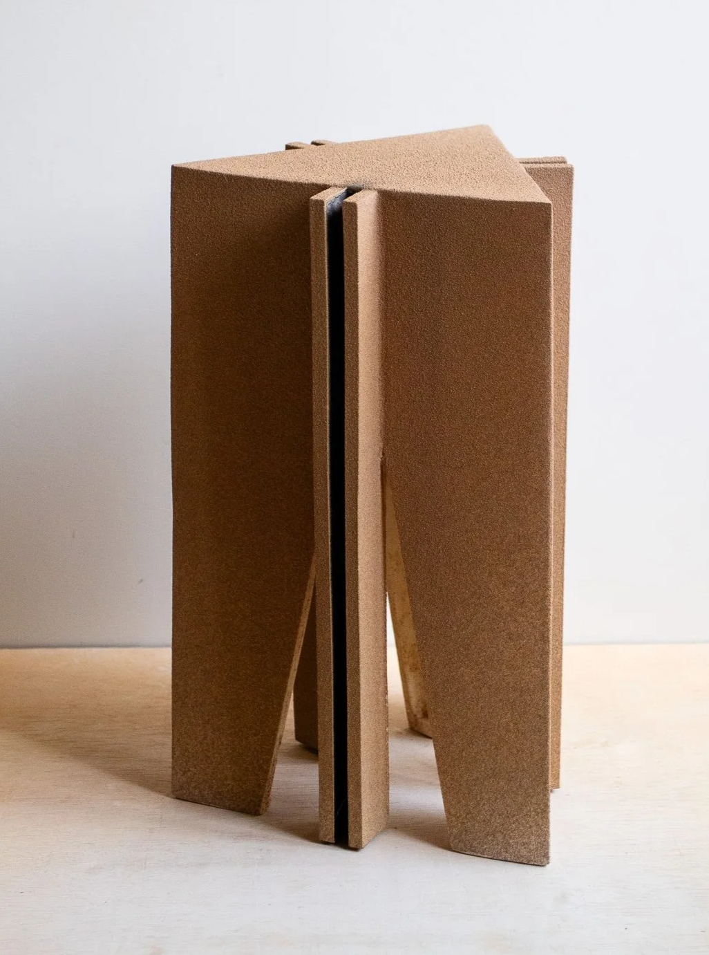 A geometric sculpture made of tan foam or cardboard with angular shapes, standing on a light wooden surface against a white background.