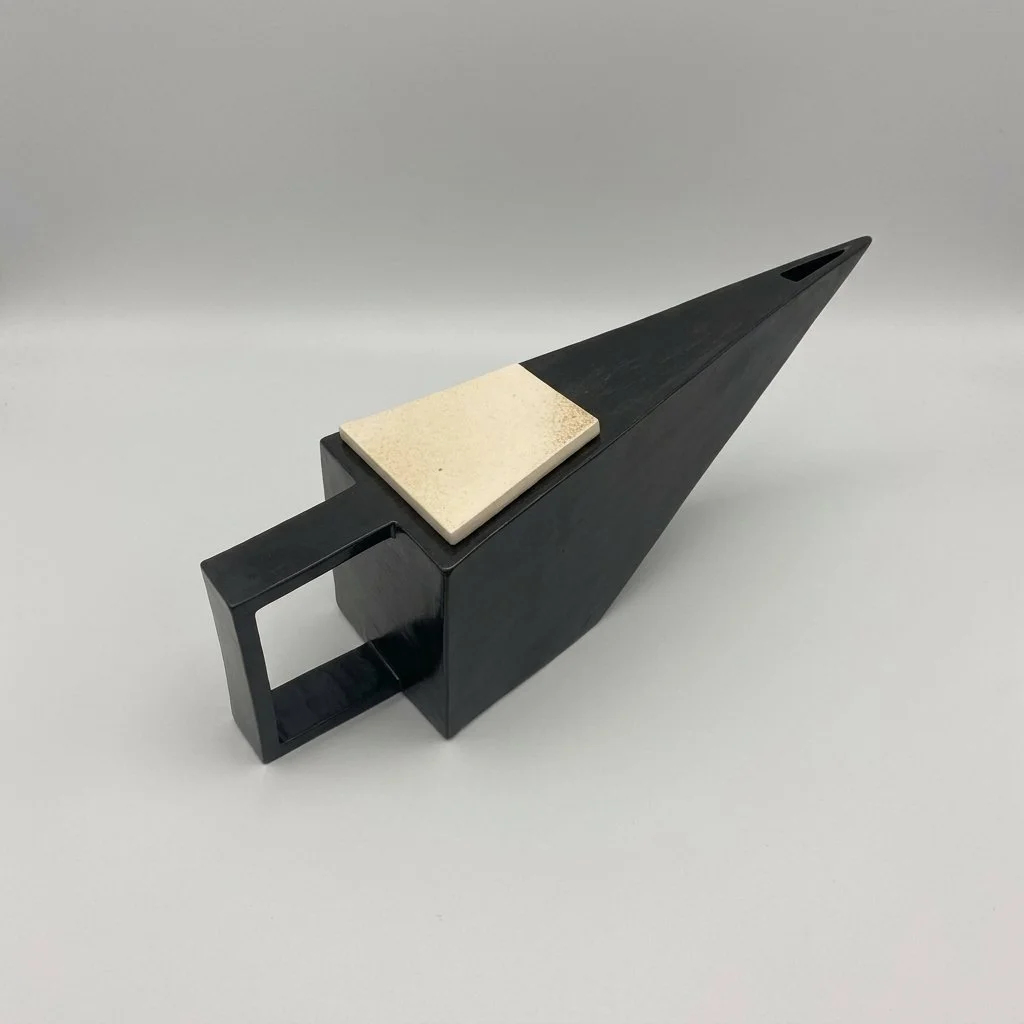 A black, geometric, arrow-shaped object with a beige rectangular piece on top, set against a gray background.