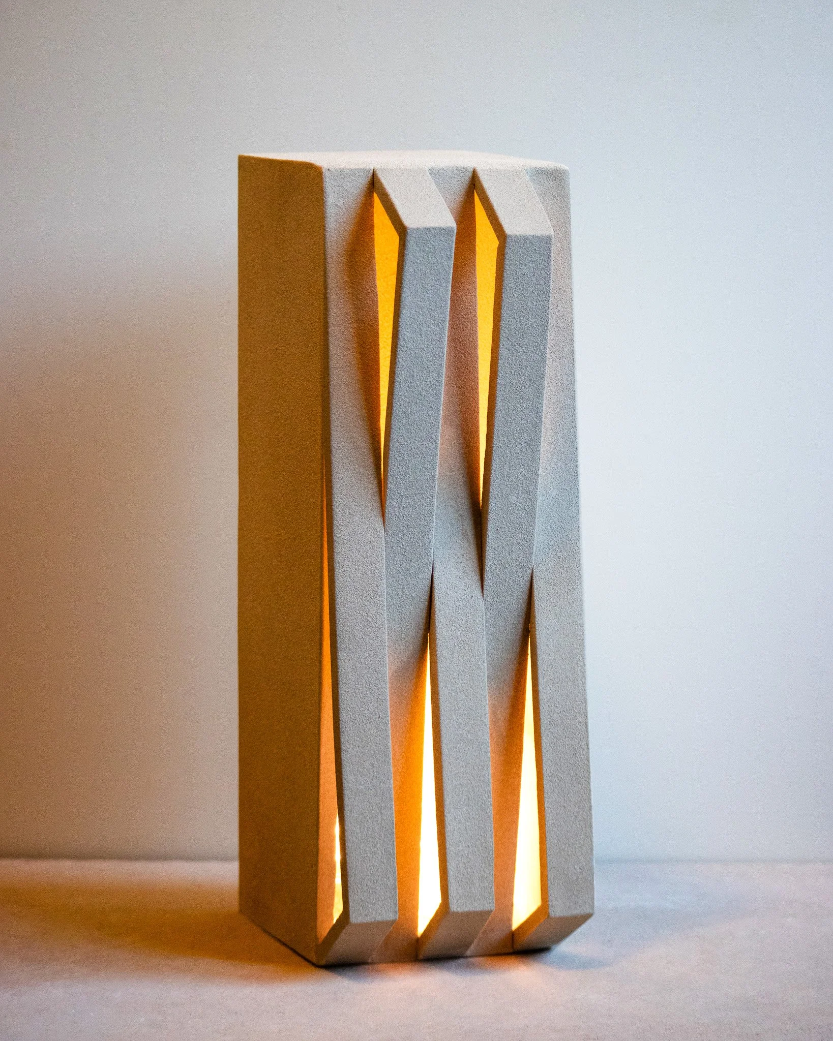 A modern decorative lamp with vertical rectangular panels made of textured white material, with orange light glowing from gaps between the panels, placed on a flat surface against a plain background.
