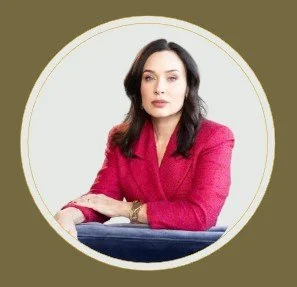 A woman with dark hair wearing a pink blazer, sitting with her arms crossed against a white background with a gold border.