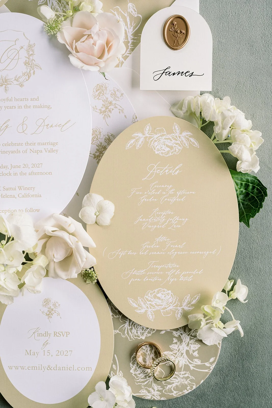 Wedding invitation suite with floral decorations and wedding rings. The suite includes a circular invitation and a beige RSVP card, all decorated with white floral designs and gold accents. There are white and pale pink flowers surrounding the cards, and a small white card with the name 'James' written in cursive and a gold wax seal.