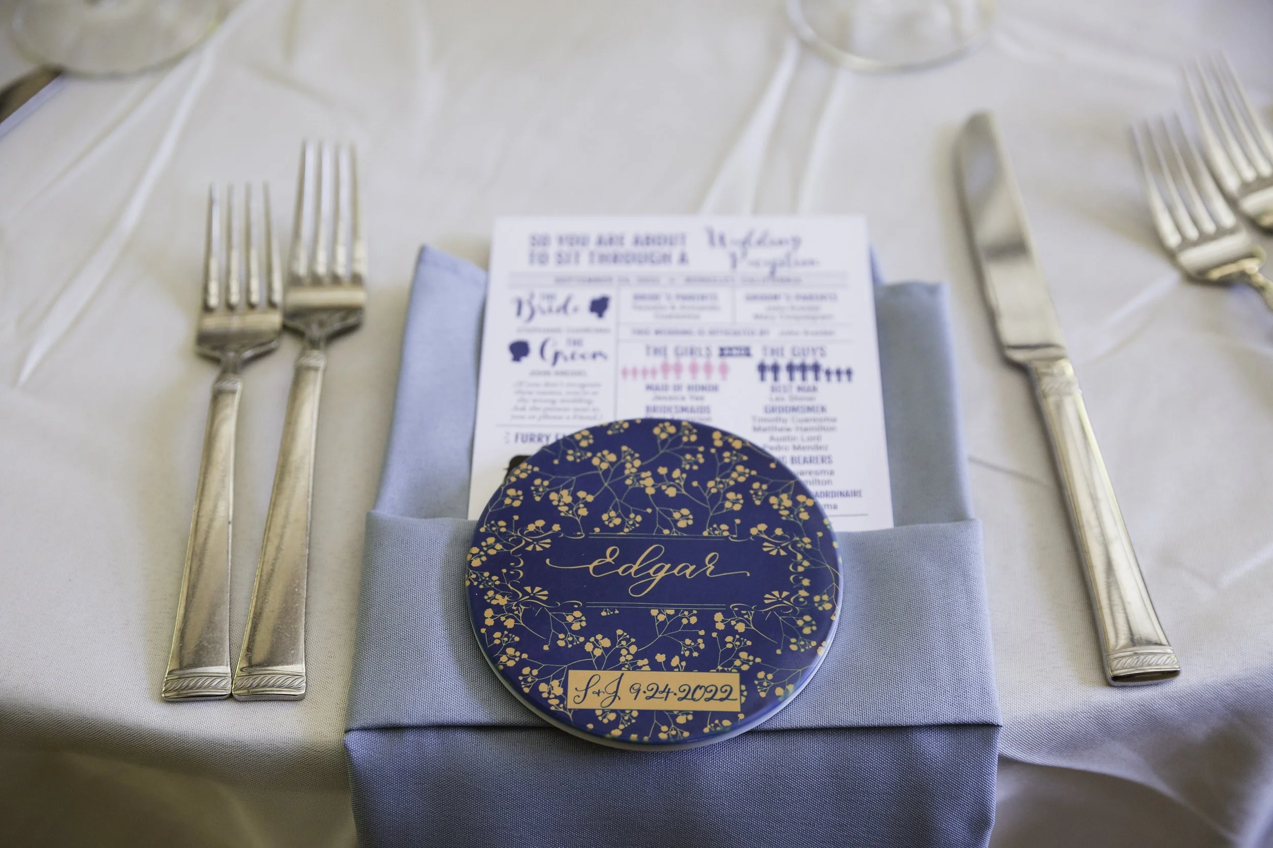 Elegant table setting with silverware and a blue napkin, featuring a round navy blue floral coaster with gold accents along with a menu in the background.