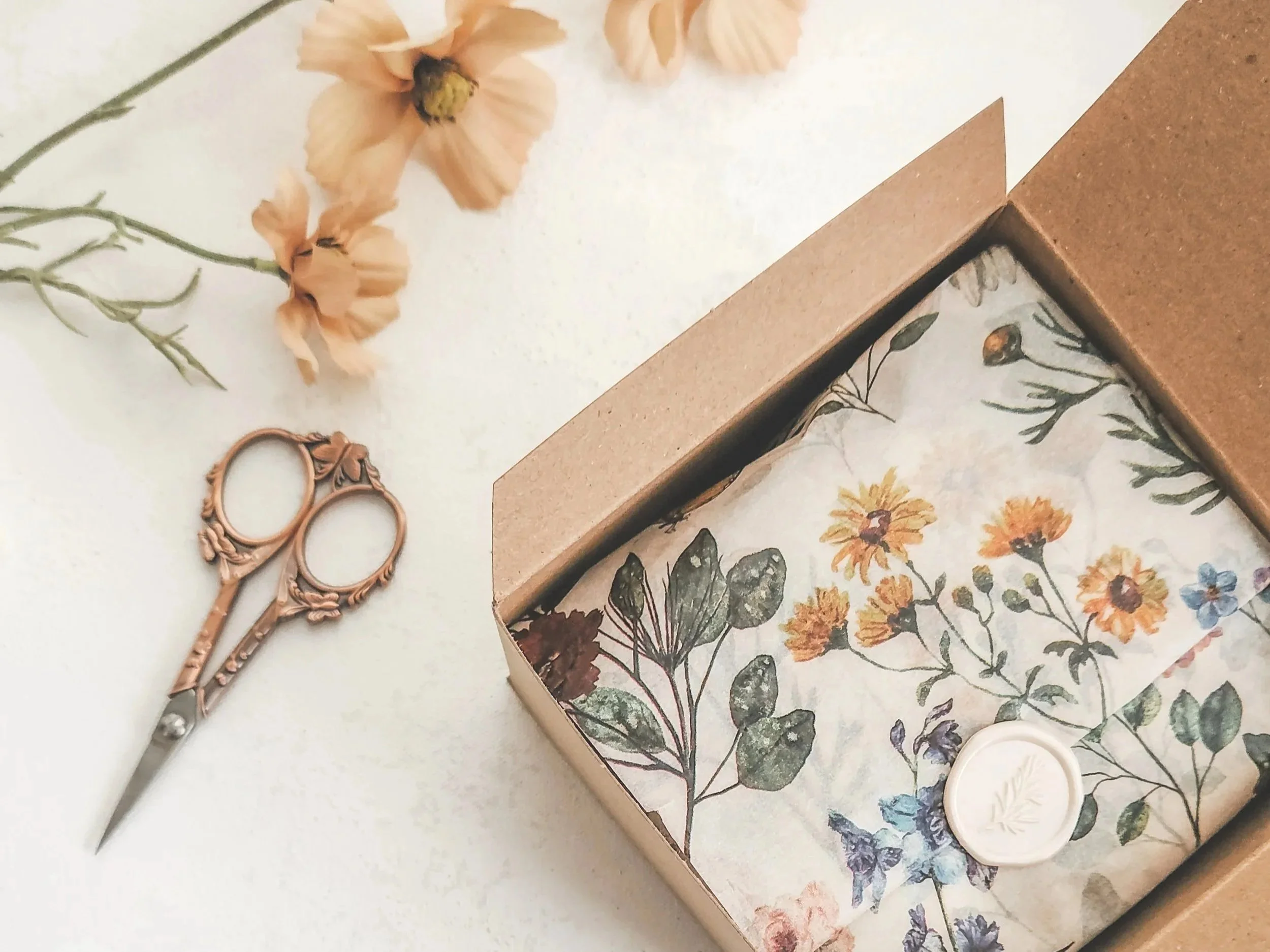 Open cardboard box containing floral-patterned paper and a wax seal with a leaf design. Nearby, there are dried flowers and ornate pink-handled scissors on a white surface.