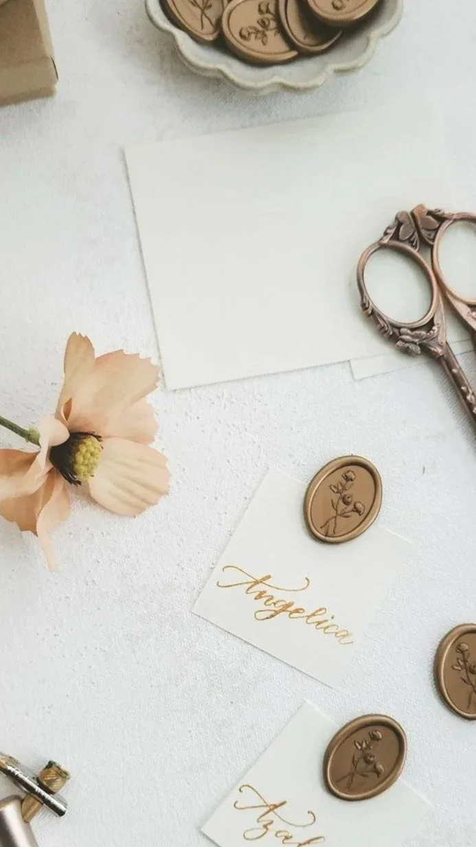 Decorative stationery, pink flower, vintage scissors, wax seals, and a sealing tool on a white surface, possibly for wedding or event invitations.
