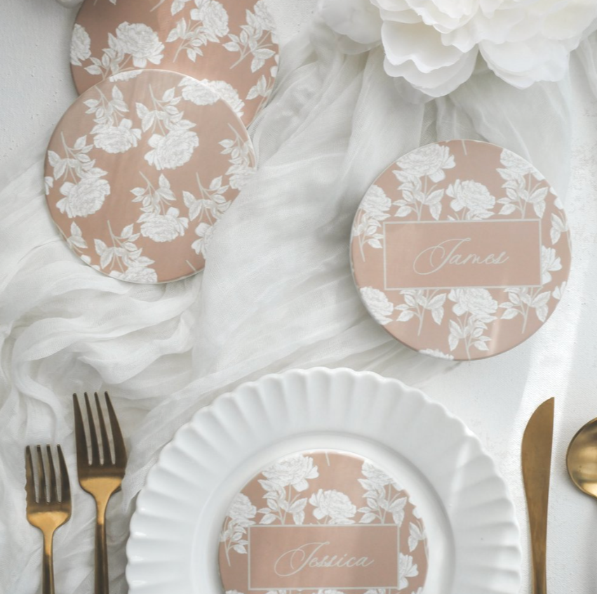 Elegant table setting with cream-colored plate, gold utensils, floral-patterned name cards, and blush-colored drink coasters with white floral designs, arranged on white fabric with a white flower accent.