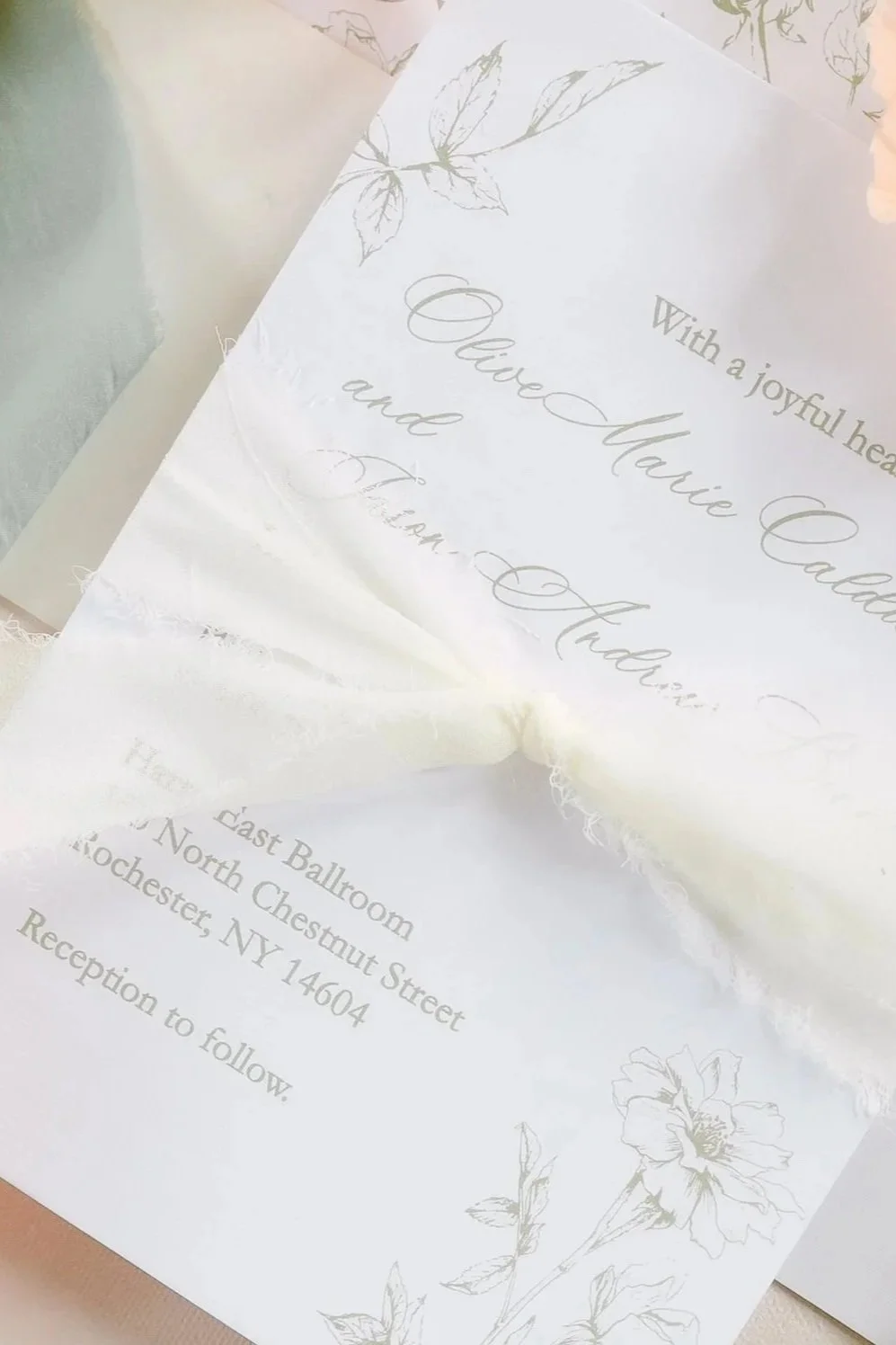 Open wedding invitation with floral border and elegant script, decorated with peach poppy flowers and a piece of white fabric.