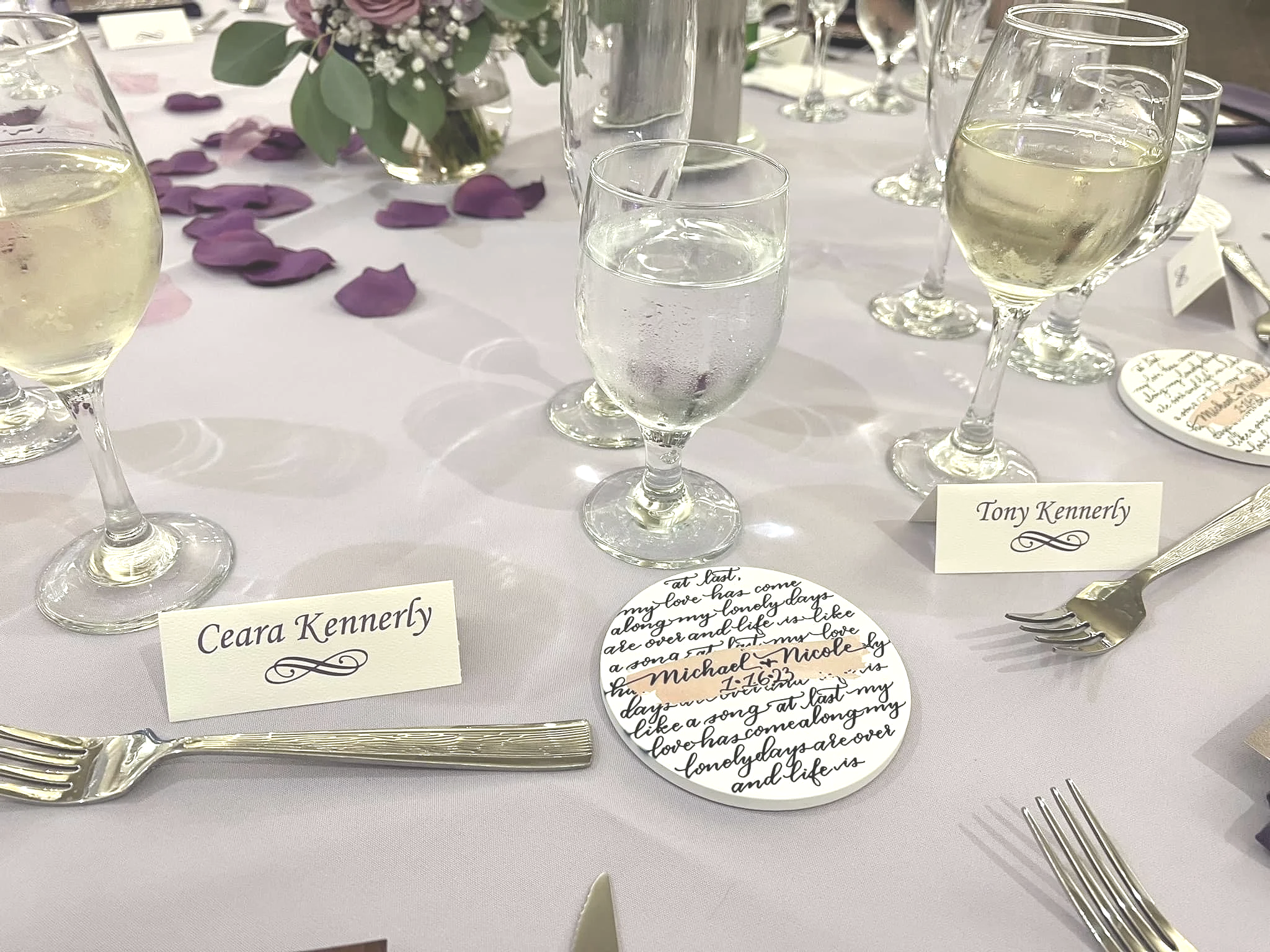 Elegant dining table setting with glasses of white wine or champagne, name cards reading 'Ceara Kennerly' and 'Tony Kennerly,' and a decorative round mirror with handwritten calligraphy. Purple rose petals and a floral centerpiece adorn the table.