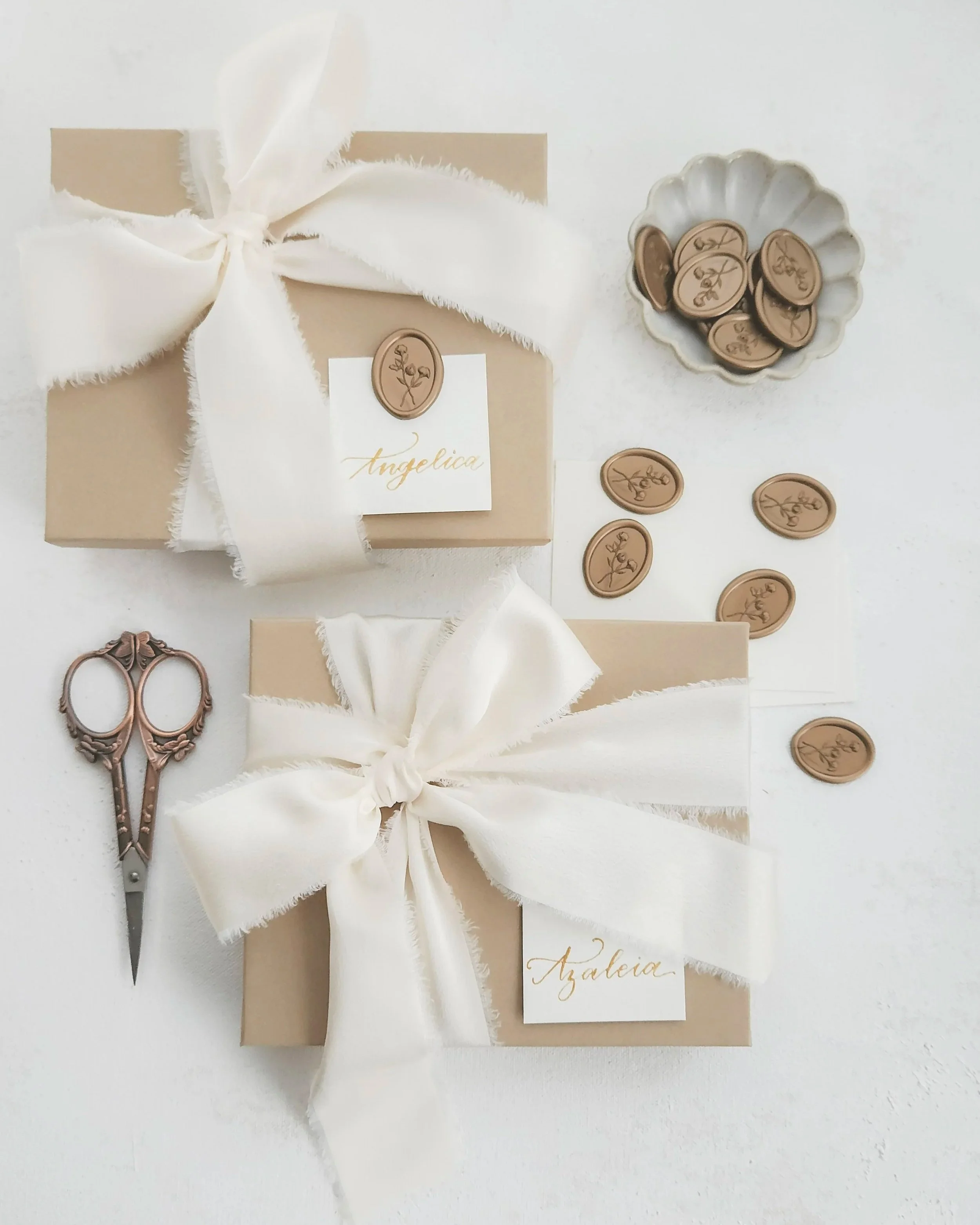 A white box wrapped with a white ribbon bow, decorated with wax seals bearing a floral design, with a pair of decorative scissors, and a white card with gold script reading 'Angelica' on a white background.