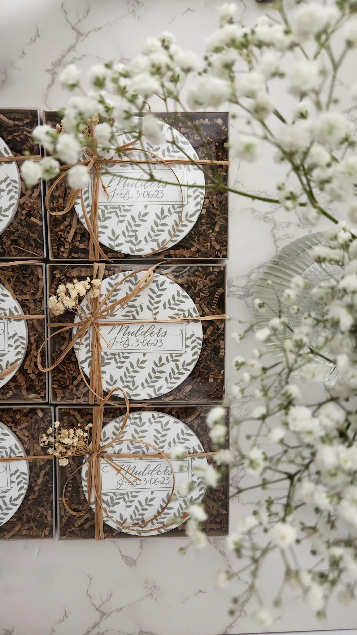 Gift boxes with round labels decorated with green leaf patterns and tied with raffia ribbons, arranged on a white marble surface, surrounded by white baby's breath flowers.