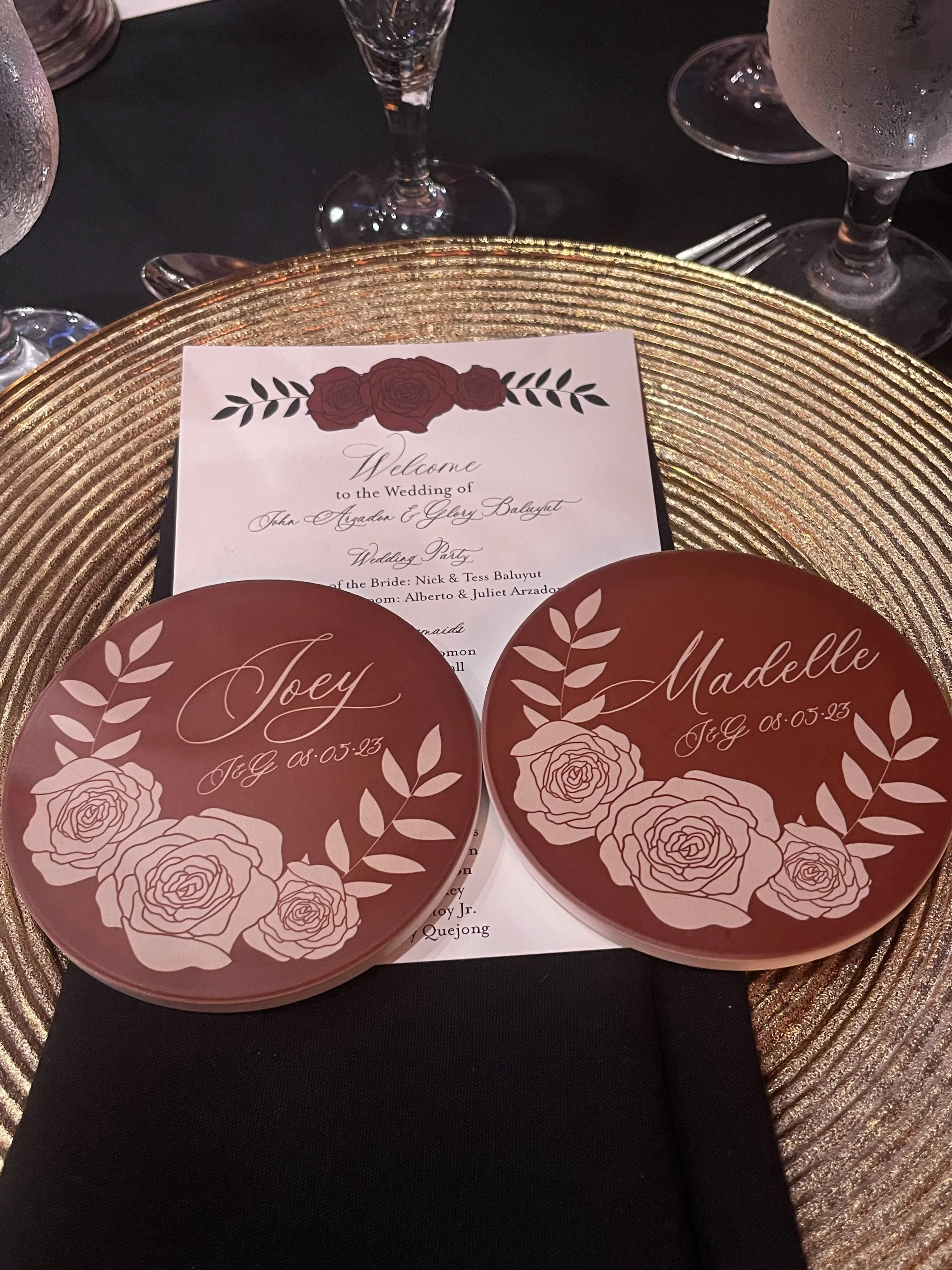 Wedding place setting with two round cards and a printed wedding program. The round with white floral designs, names 'Joey' and 'Madeleine', and the date '08-05-23' written on them. The wedding program is behind the cards, with a floral header and cursive text welcoming guests to the wedding of John Ayden and Glozy Balyut.