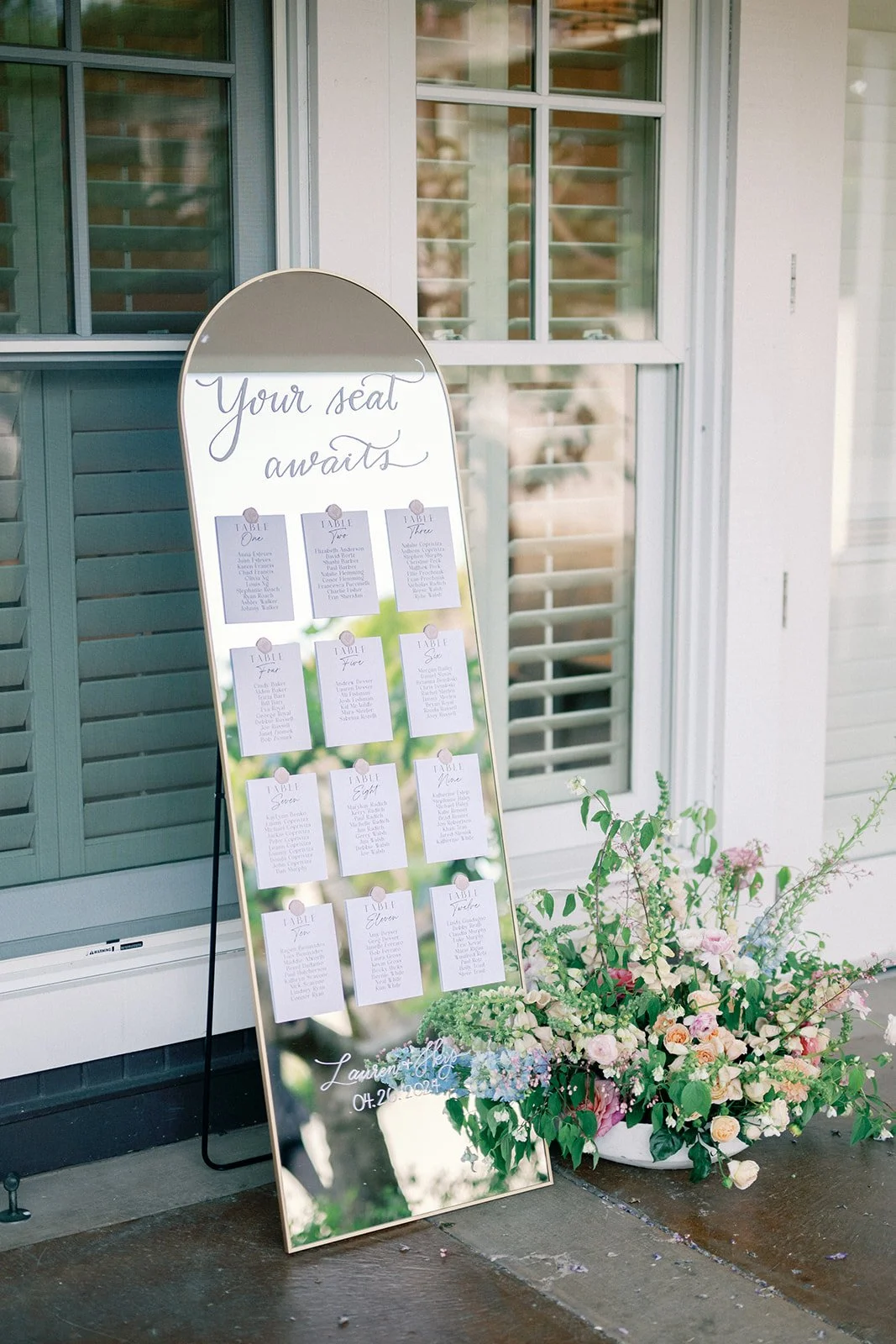 A wedding seating chart displayed on a tall, oval mirror with the words 'Your seat awaits' at the top. The chart has individual pages for each table with guest names. Next to the mirror is a large bouquet of flowers on a concrete surface.