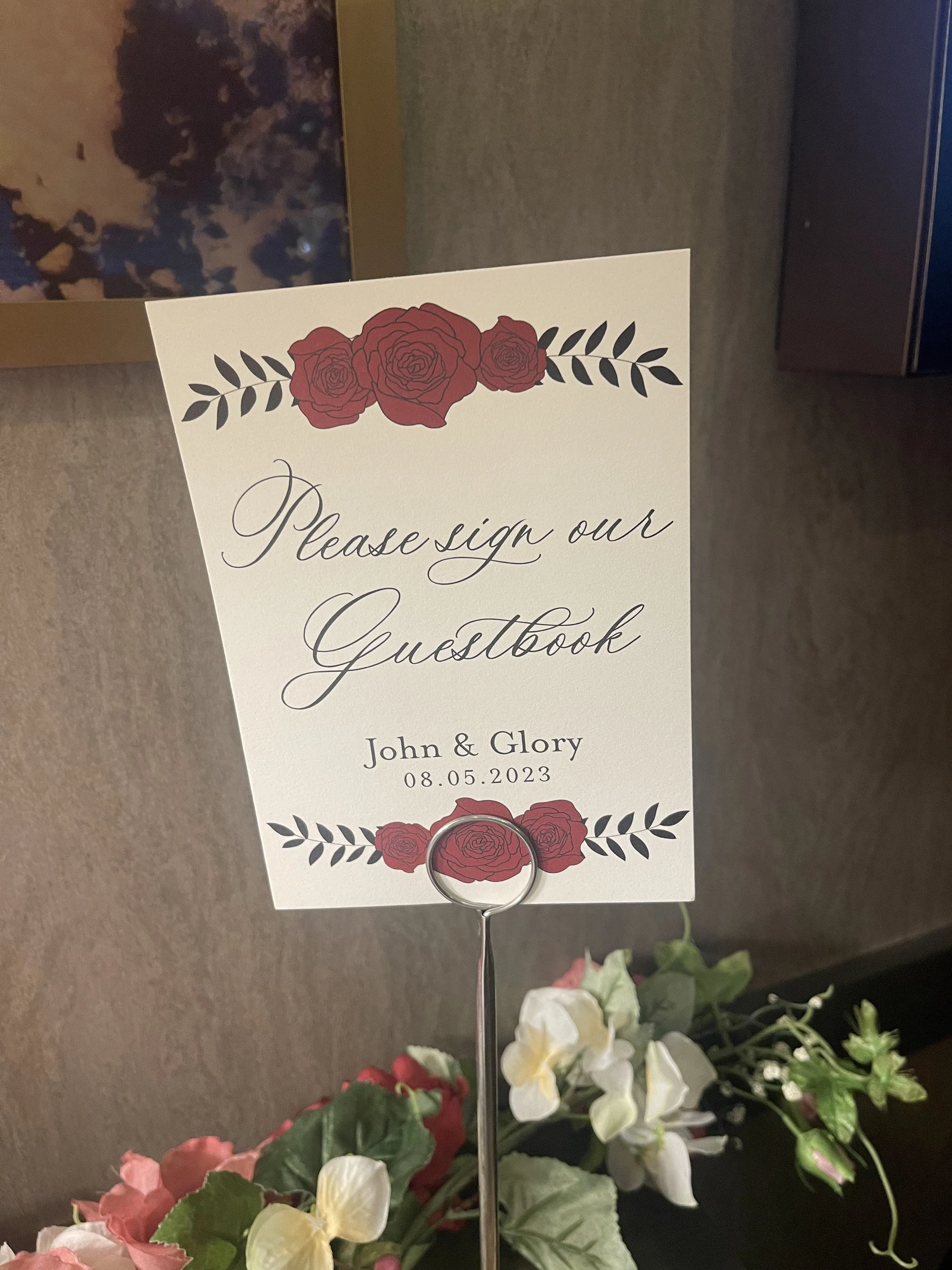 Sign in a wedding guestbook area with red and white flowers, instructing guests to sign the guestbook for John and Glory on August 5, 2023.