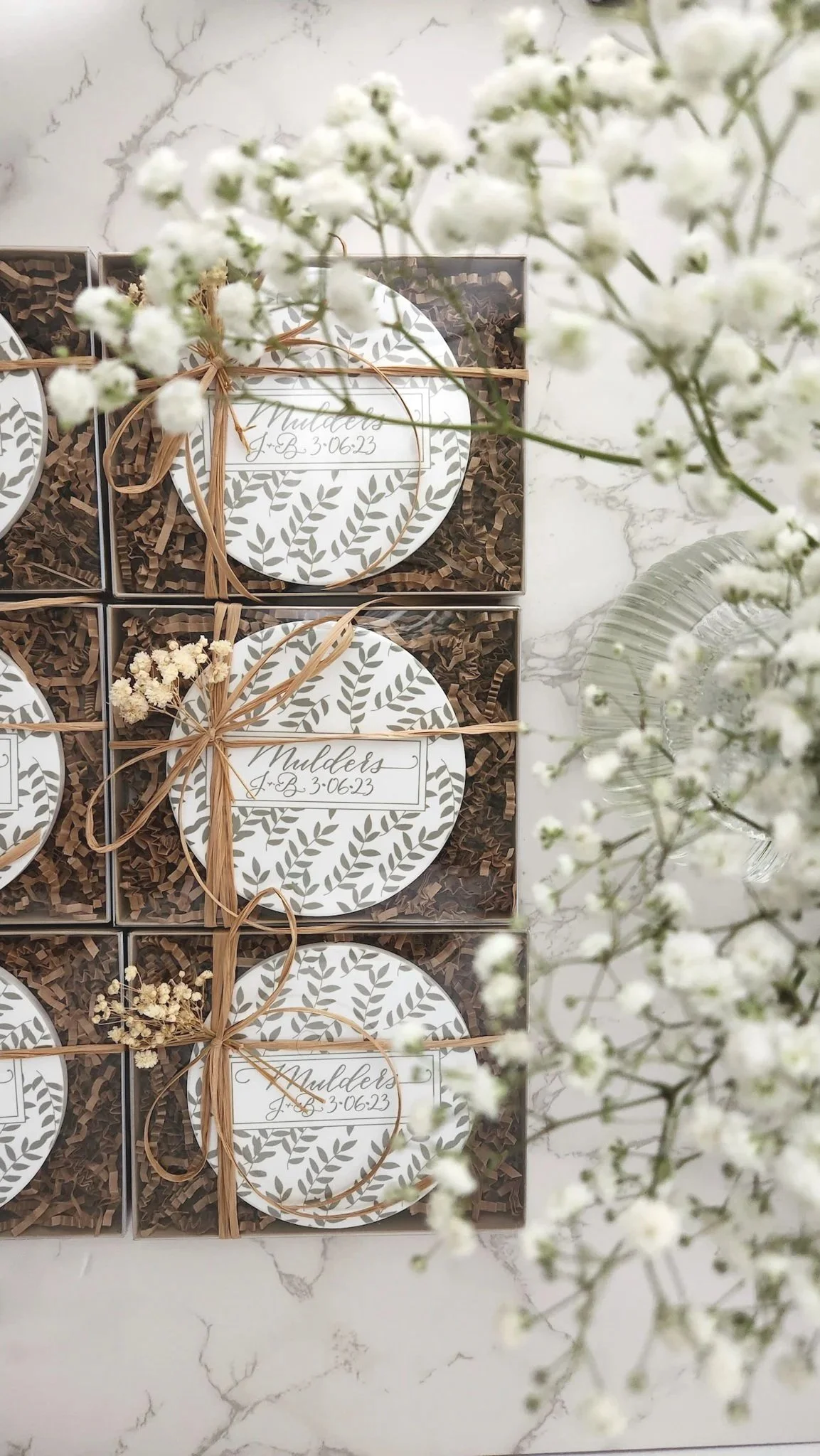 Gift boxes with round labels decorated with green leaf patterns and tied with raffia ribbons, arranged on a white marble surface, surrounded by white baby's breath flowers.