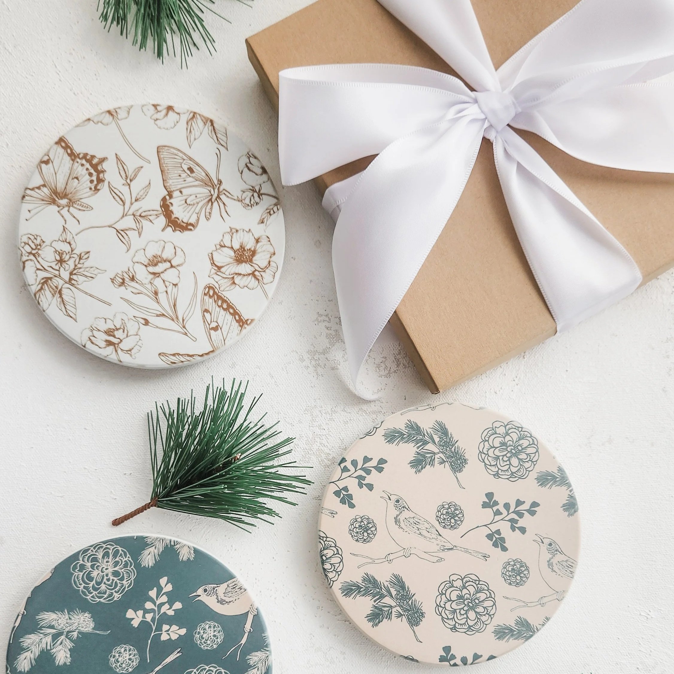 Decorative coasters with floral and bird designs, some with butterflies, arranged on a white surface with greenery.