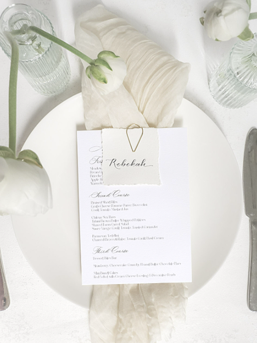A elegant wedding place setting with a white plate, a menu card labeled 'Rebekah,' a napkin, a cream-colored cloth, and floral decorations including white roses in glass vases.
