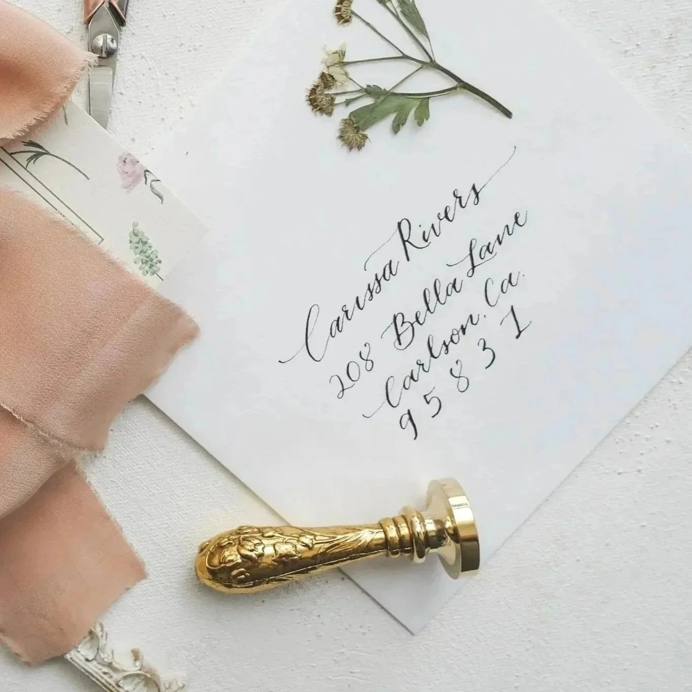 A white envelope with elegant black handwritten address, surrounded by dried flowers, a cream-colored fabric, and a gold decorative object.