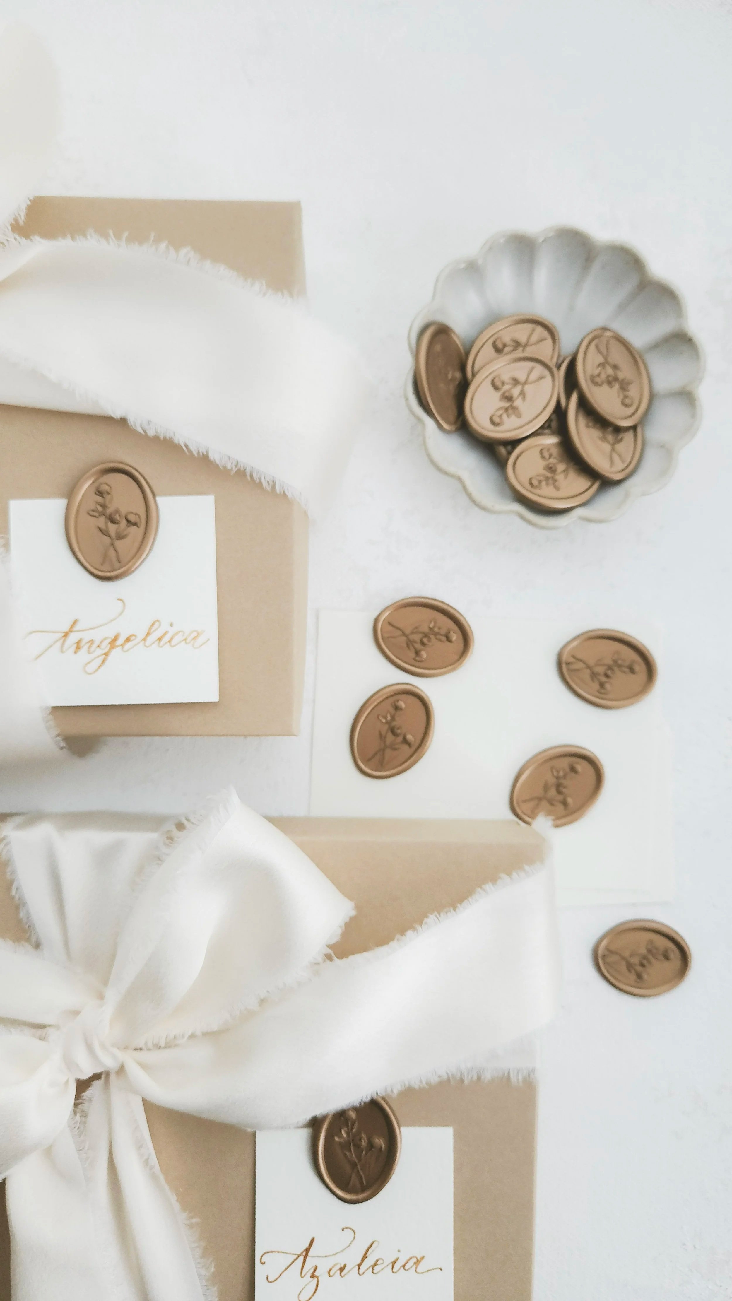 Beige gift boxes decorated with white ribbons, with wax seals featuring floral designs and the names Angelica and Azaleia, next to a bowl of wax seals.