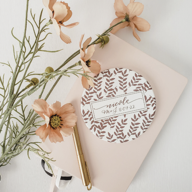A floral arrangement with light peach-colored flowers and green foliage, a cream-colored notebook with a round sticker that says 'Nicole, Mt 8:07.22' surrounded by a leaf pattern, and a gold pen on top of the notebook.