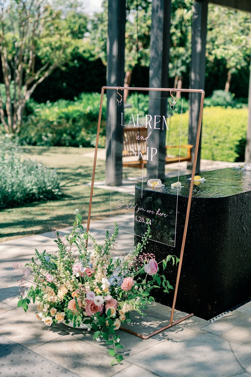 A wedding sign with the names Lauren and Skip, in an outdoor setting with trees and greenery, and a flower arrangement at the base of the sign.