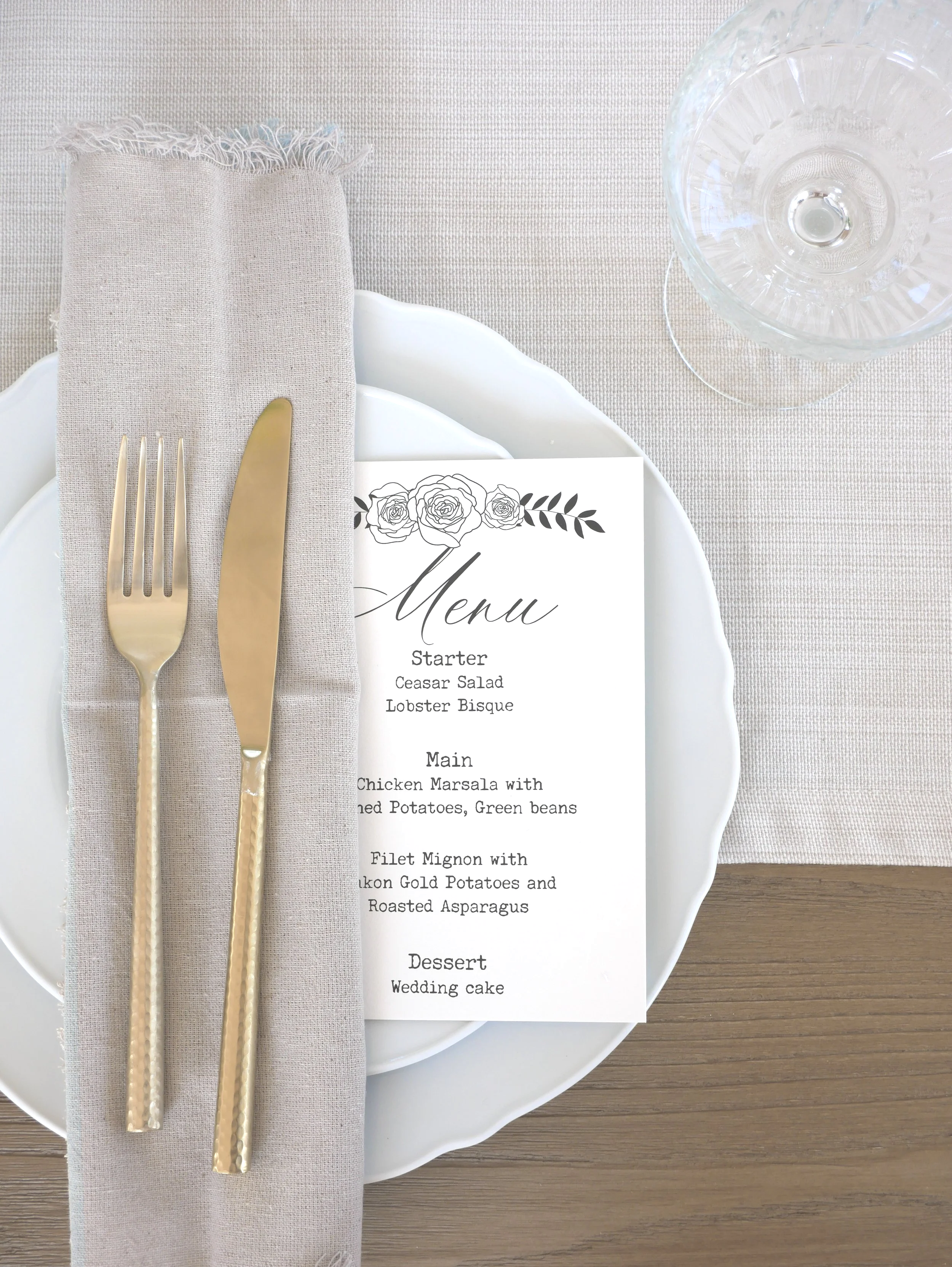 Elegant table setting with a white plate, grey napkin, gold fork and knife, a wedding menu card with floral design, clear water glass, and a wooden table surface.