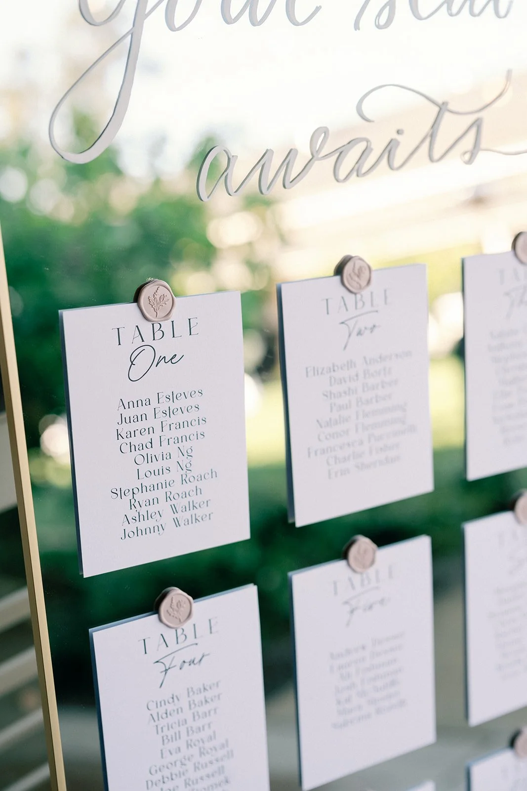 Sign with seating chart for a wedding, showing tables labeled One, Two, and Four with guest names listed on white cards, pinned with wax seal magnets.