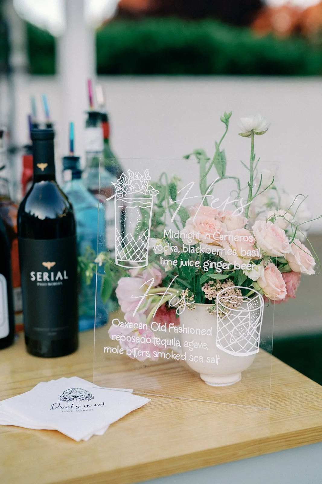 A clear acrylic menu with cocktail recipes, a white vase with pink and white roses, and bottles of alcohol in the background on a wooden bar counter.
