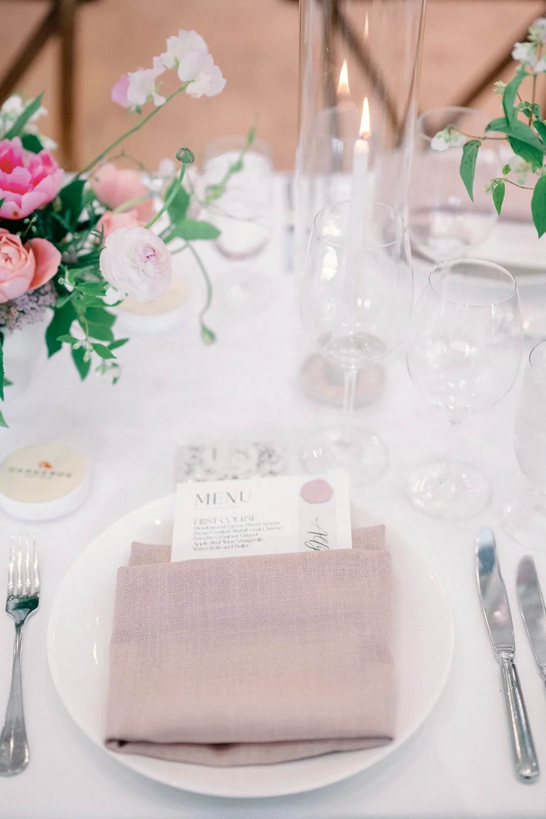 Elegant table setting with pink napkin on white plate, clear glassware, a menu card, and floral centerpiece with pink and white flowers.