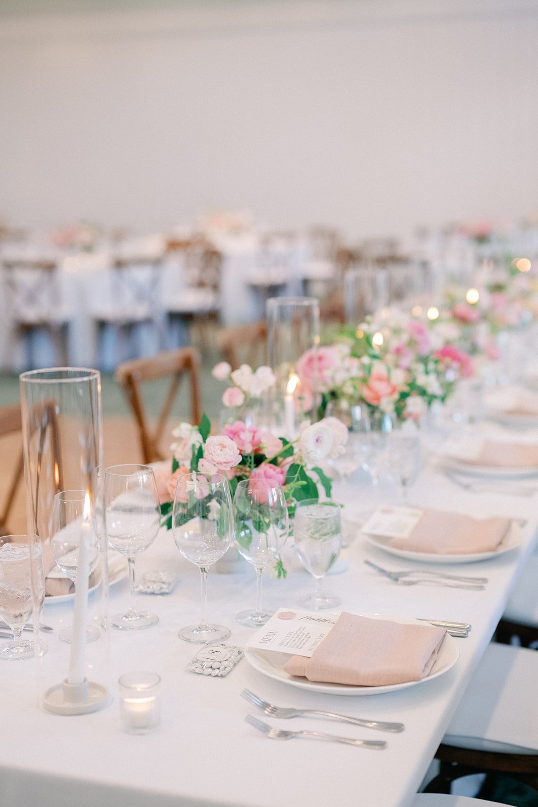 A wedding reception table decorated with pink and white flowers, candles, champagne glasses, plates, silverware, and printed menus.