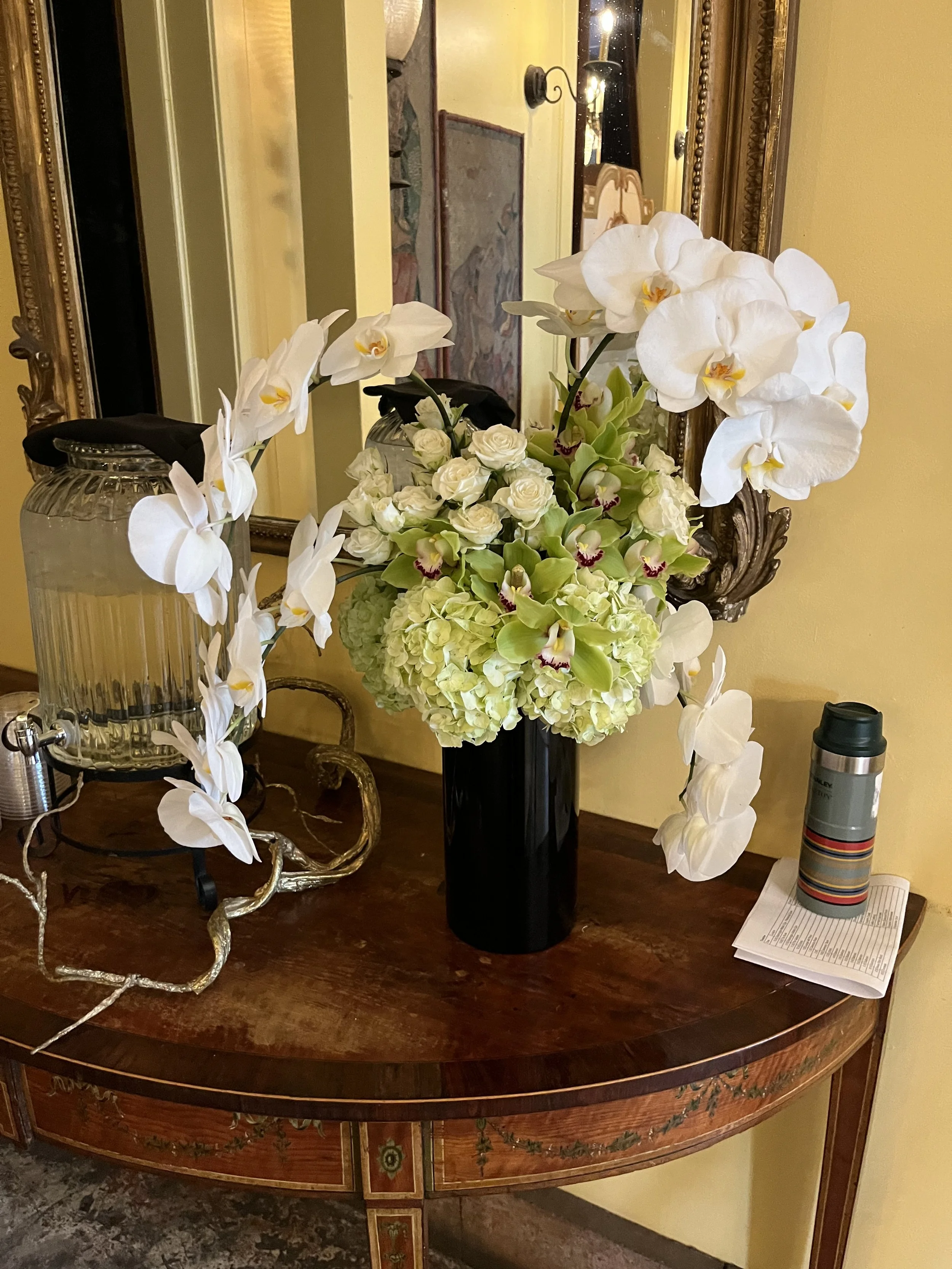 Elegant floral arrangement with white orchids and light green flowers in a black vase on a wooden table with a mirror and decorative items.