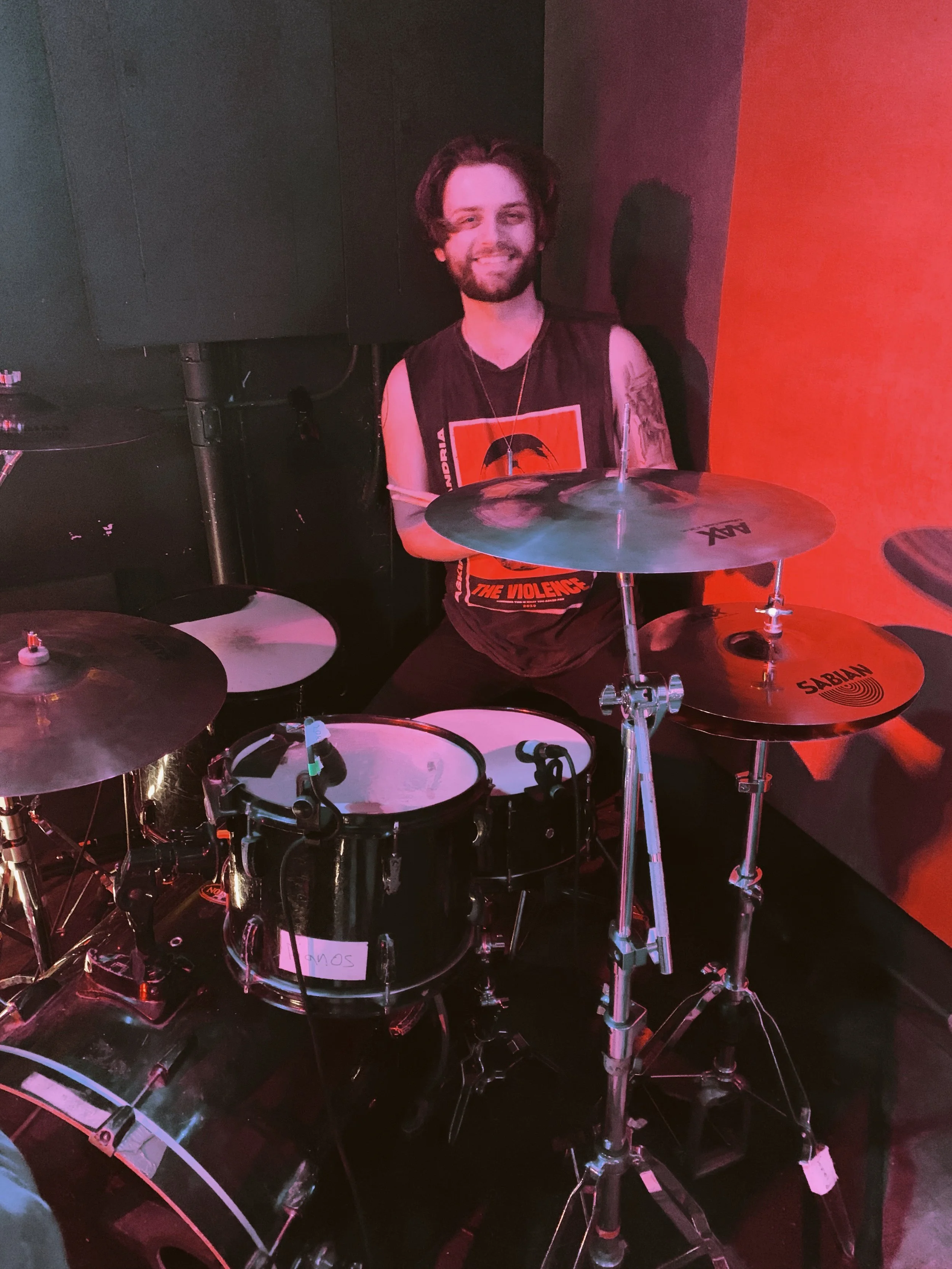 A man with long hair and a beard sitting behind a drum set, smiling at the camera in a dimly lit room with a red wall.