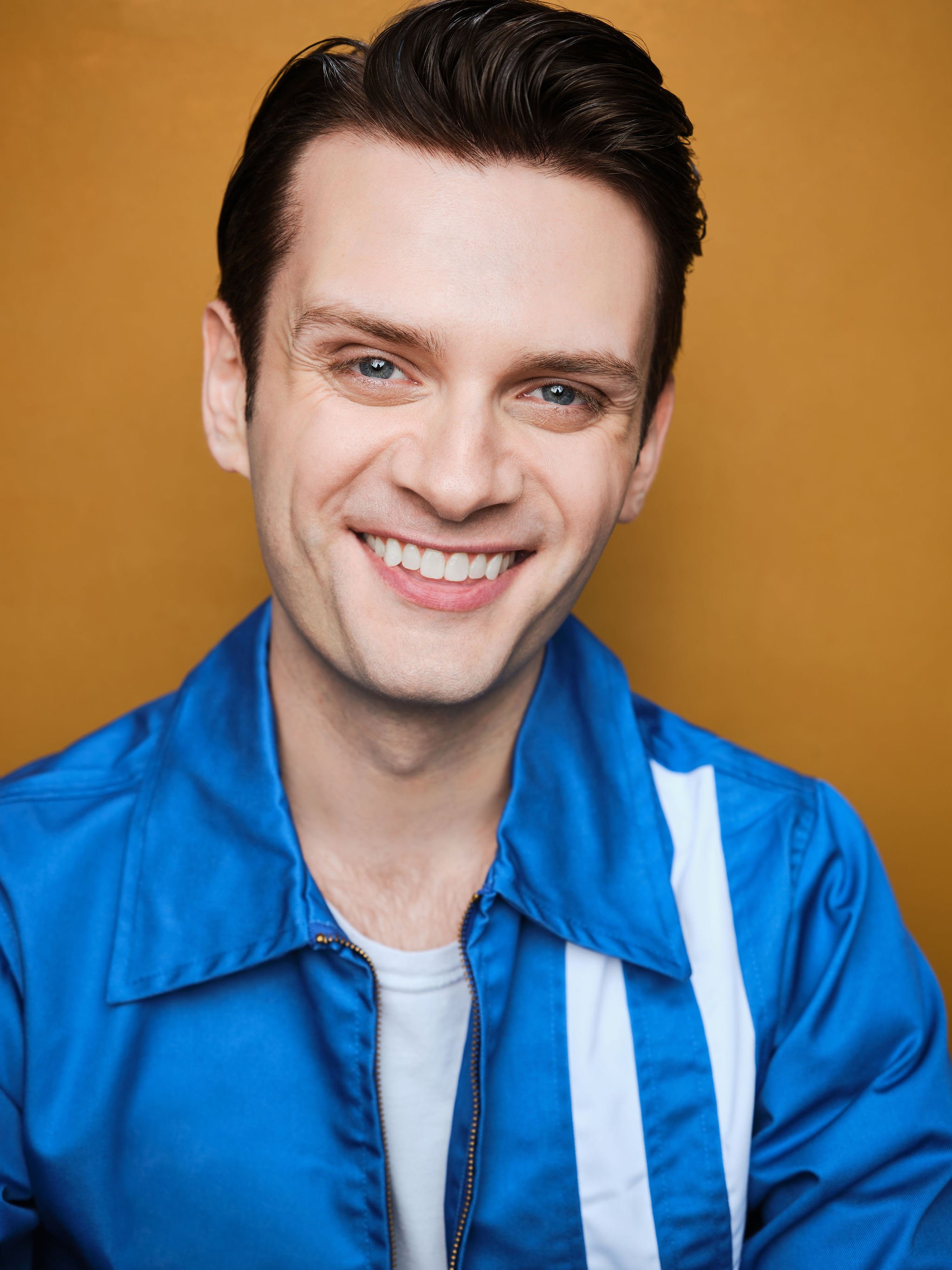 A smiling man with dark hair and blue eyes, wearing a blue jacket and a white shirt, against a yellow background.