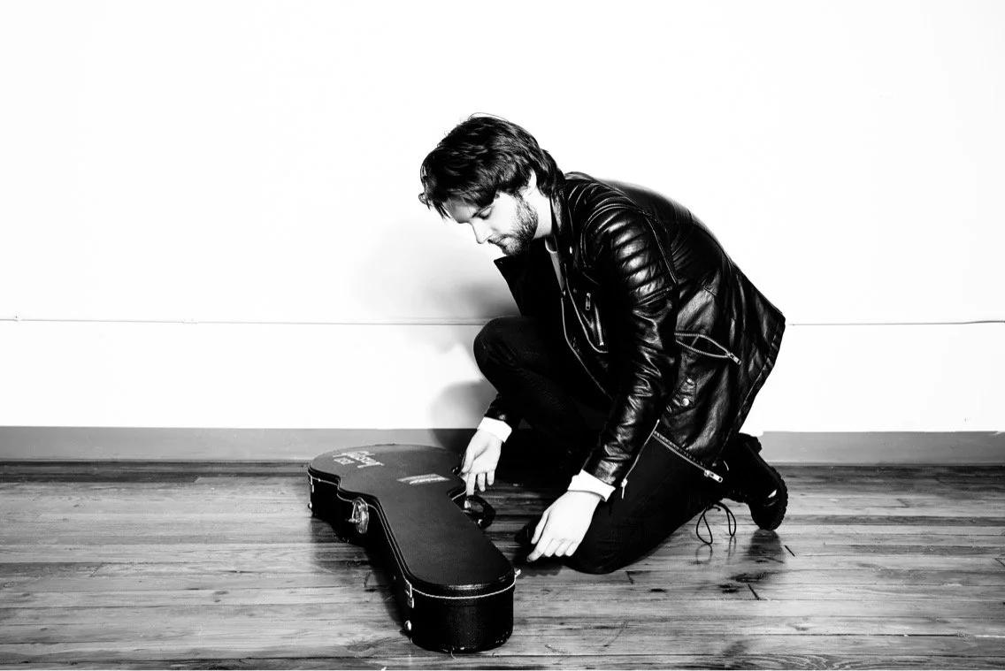 A man in a leather jacket kneeling on a wooden floor, preparing a guitar case.