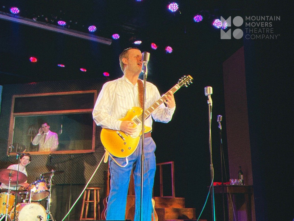 Music performance on stage with a guitarist singing into a microphone, a drummer playing drums in the background, and a soundproof window with a reflection of the guitarist, at Mountain Movers Theatre Company.